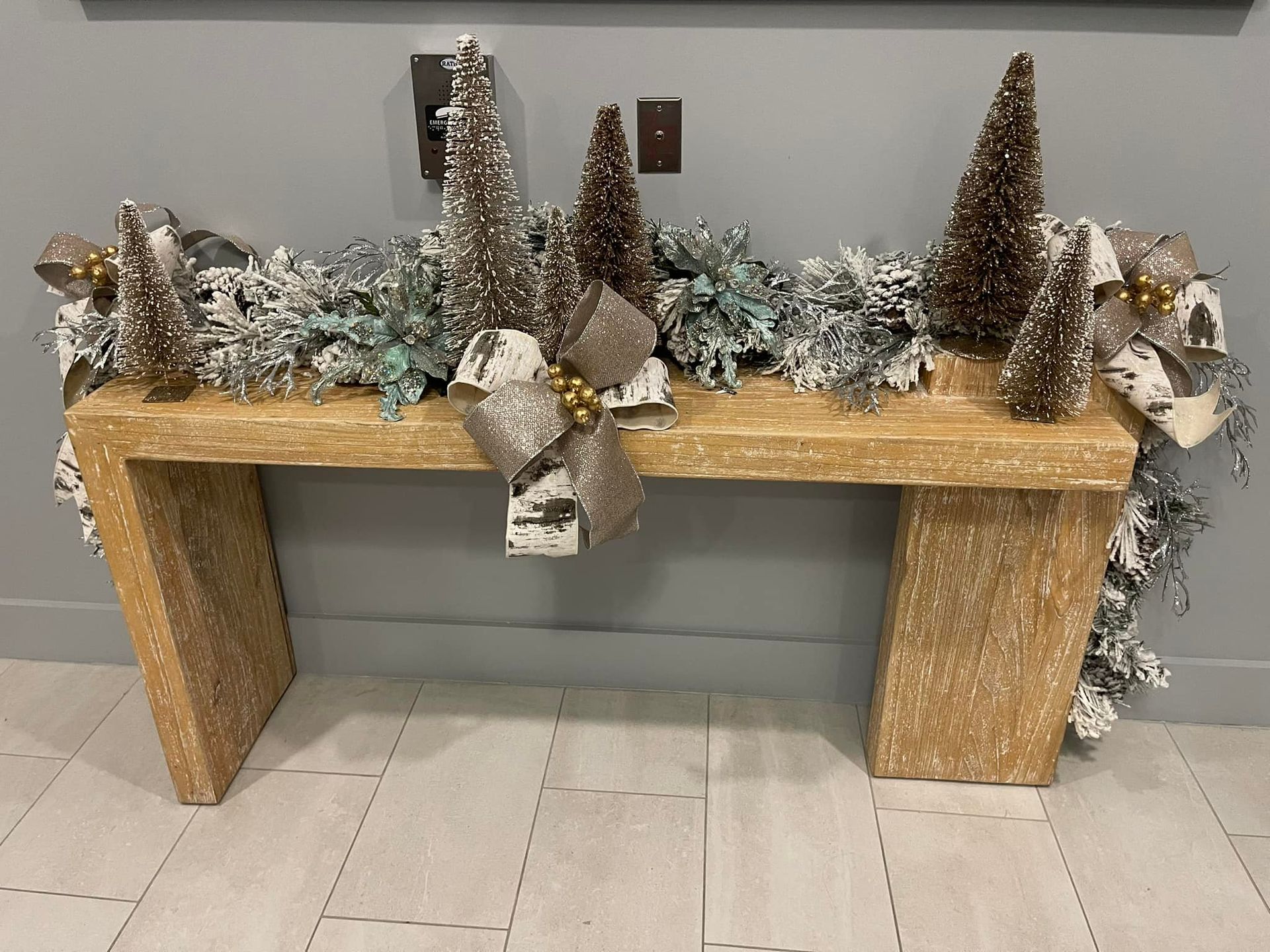 A rustic wooden console table decorated with frosted greenery, miniature trees, and bows against a grey wall.
