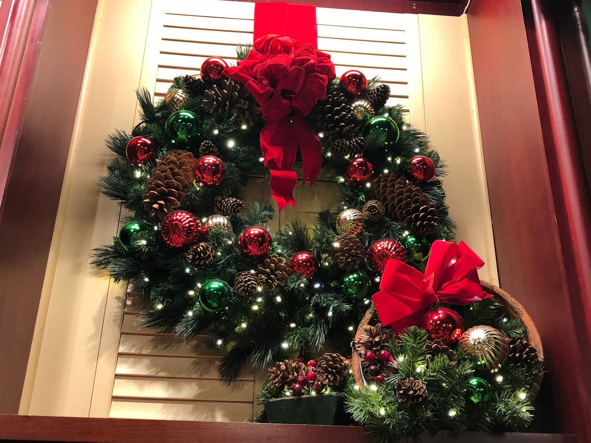 A large lit Christmas wreath with a red bow hangs above a smaller, matching basket arrangement against window blinds.