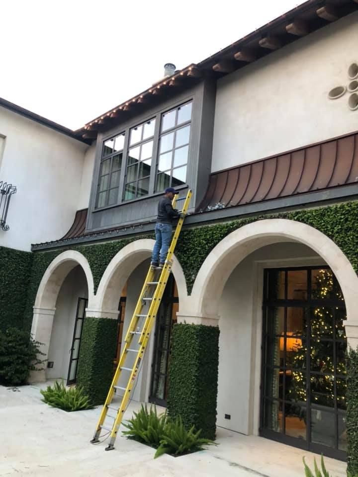A person on a tall yellow ladder cleaning the exterior window of a multi-story home with arched doorways and ivy columns.