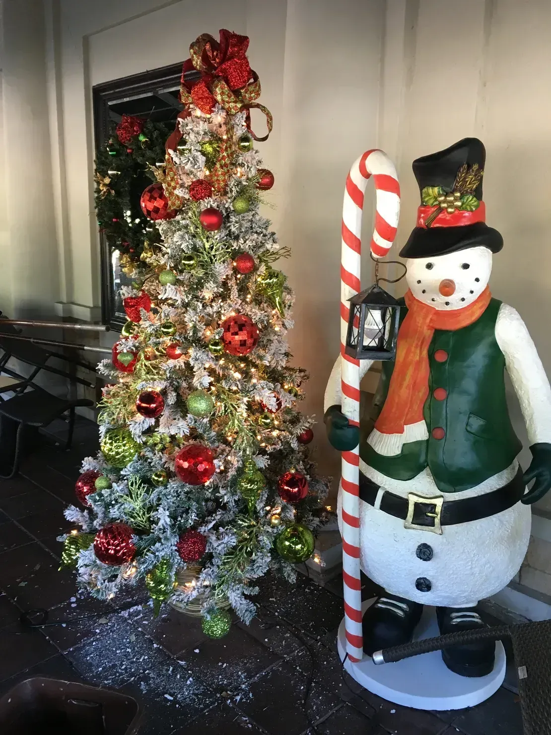 A decorated Christmas tree with red and green ornaments next to a snowman statue holding a candy cane lantern.
