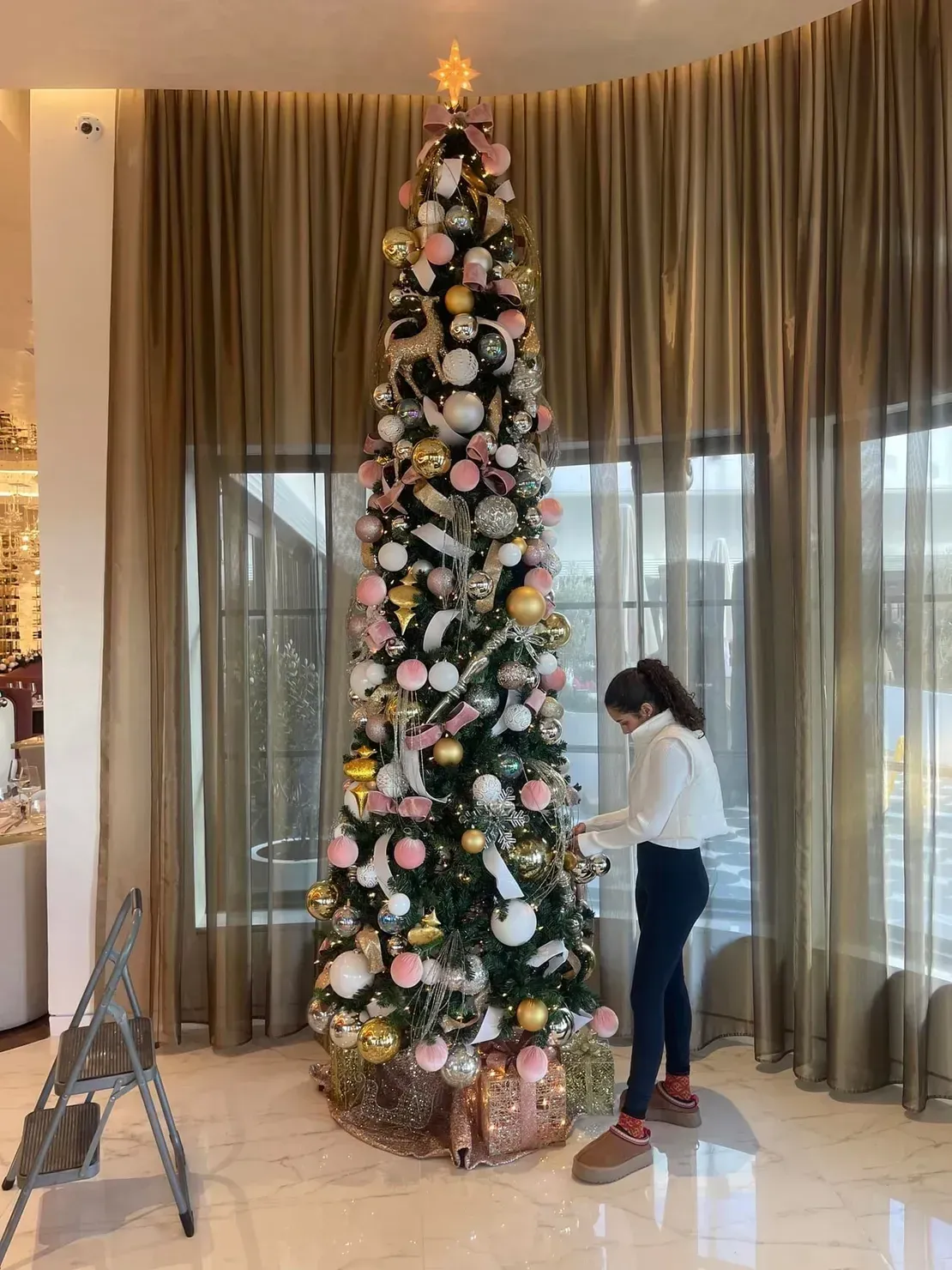 A person decorating a tall, slim Christmas tree adorned with gold, white, and pink ornaments in a room with draped curtains.