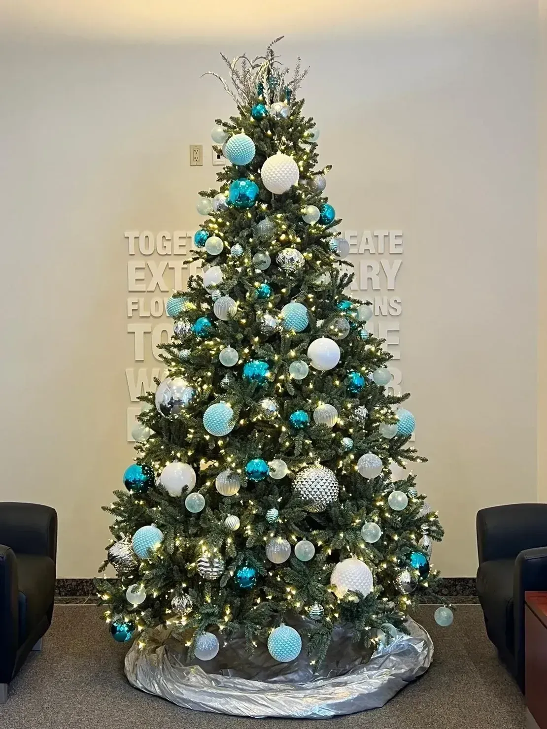 A decorated Christmas tree with teal, white, and silver ornaments in a room with two chairs and a wall sign.