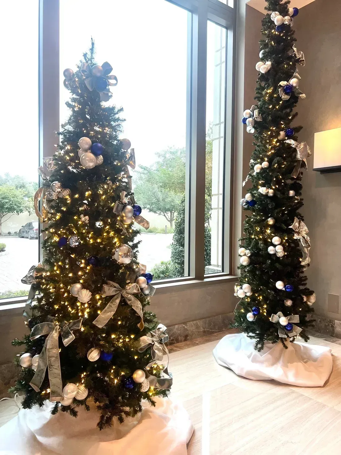 Two decorated Christmas trees stand on white tree skirts inside a room with a large window.
