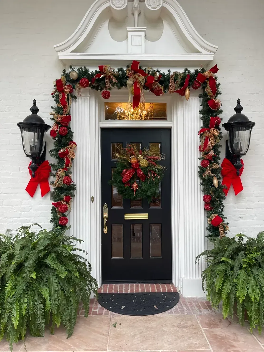 A black front door decorated for Christmas with a wreath, a garland with red bows, and potted ferns on both sides.