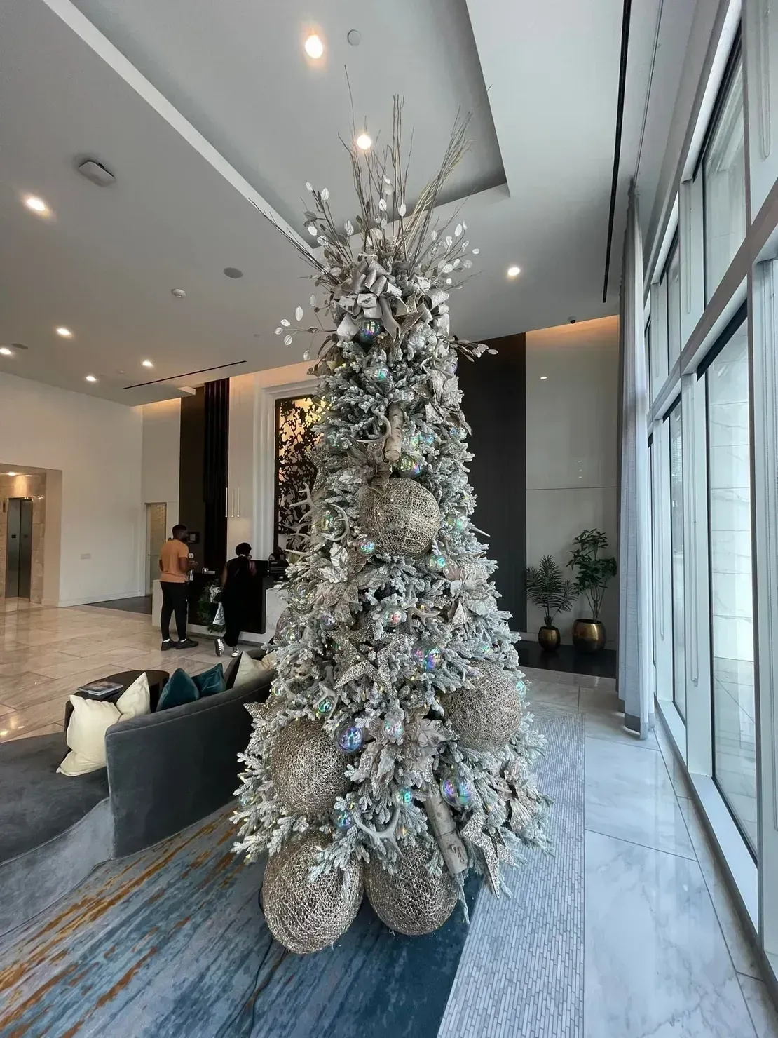 A tall, silver-decorated Christmas tree stands in a modern building lobby with marble floors and floor-to-ceiling windows.