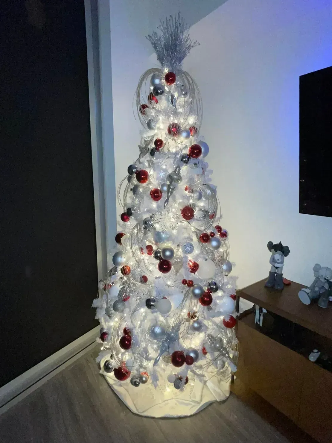 A white Christmas tree decorated with red and silver baubles and glowing lights, standing in a corner of a room.