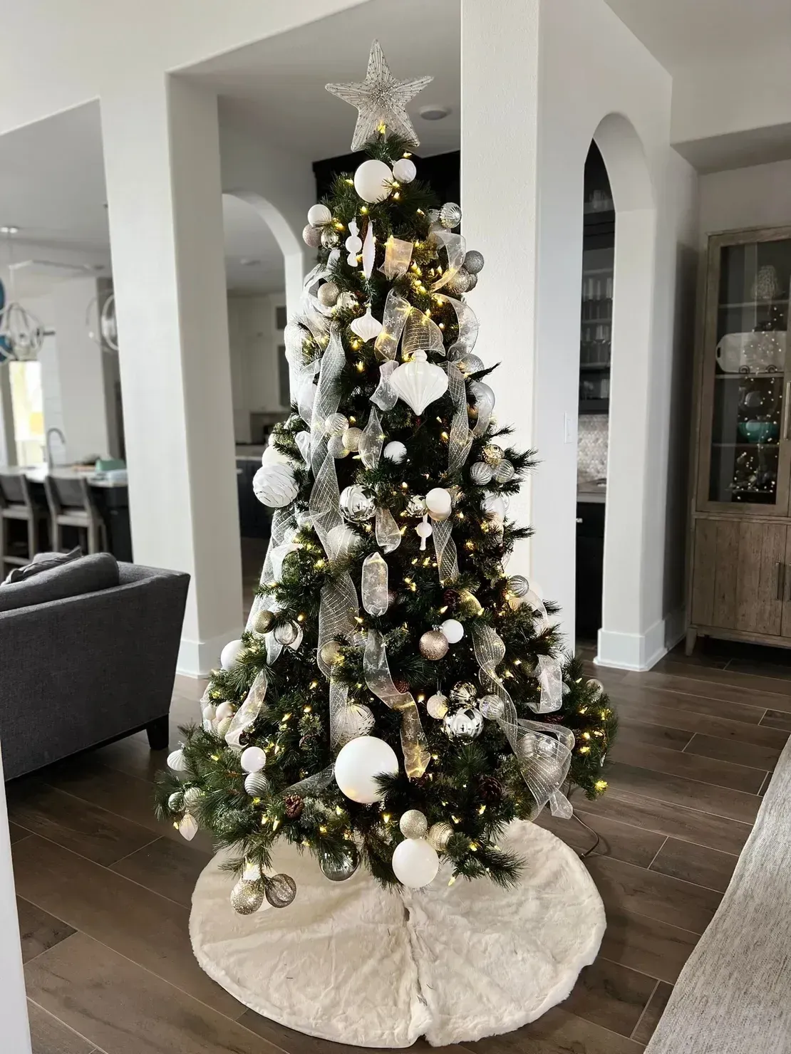 A decorated Christmas tree with white and silver ornaments, ribbon, and a star topper, set on a white fur tree skirt.