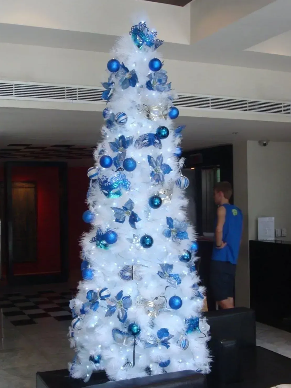 A tall white artificial Christmas tree decorated with blue ornaments and silver accents stands in an indoor lobby.