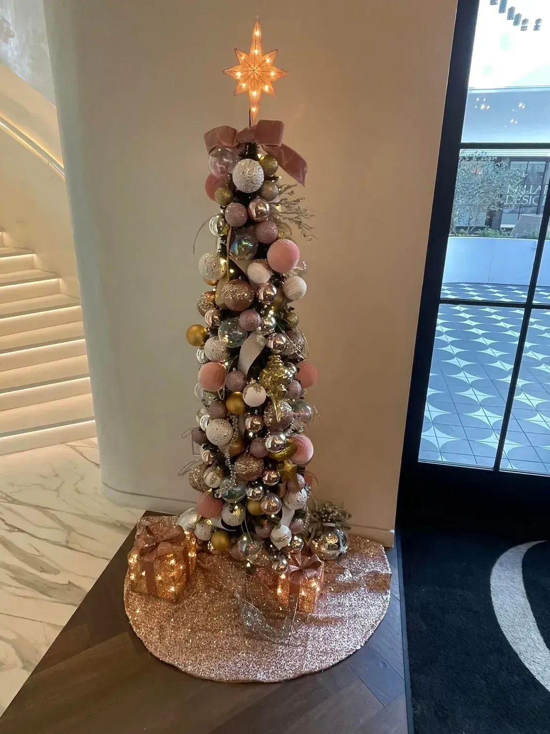 A narrow, slim Christmas tree decorated with pink, gold, and white ornaments, topped with a star, on a sequined tree skirt.