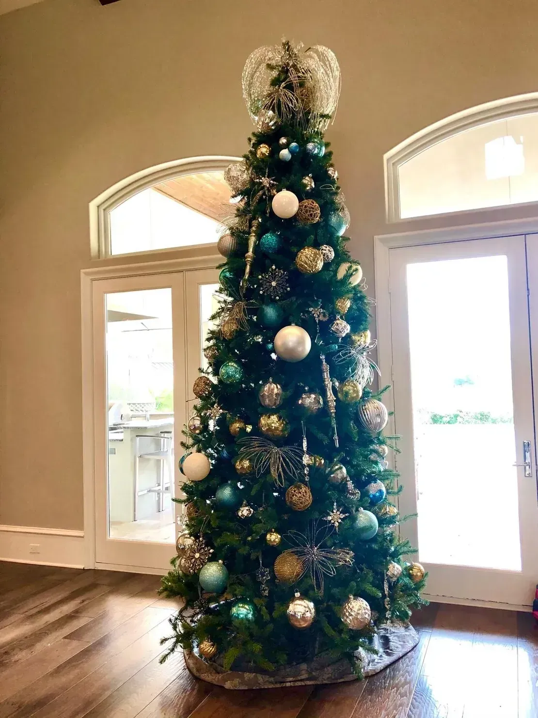 A tall, decorated Christmas tree with blue, silver, and gold ornaments, topped with a sparkling star, in a bright room.