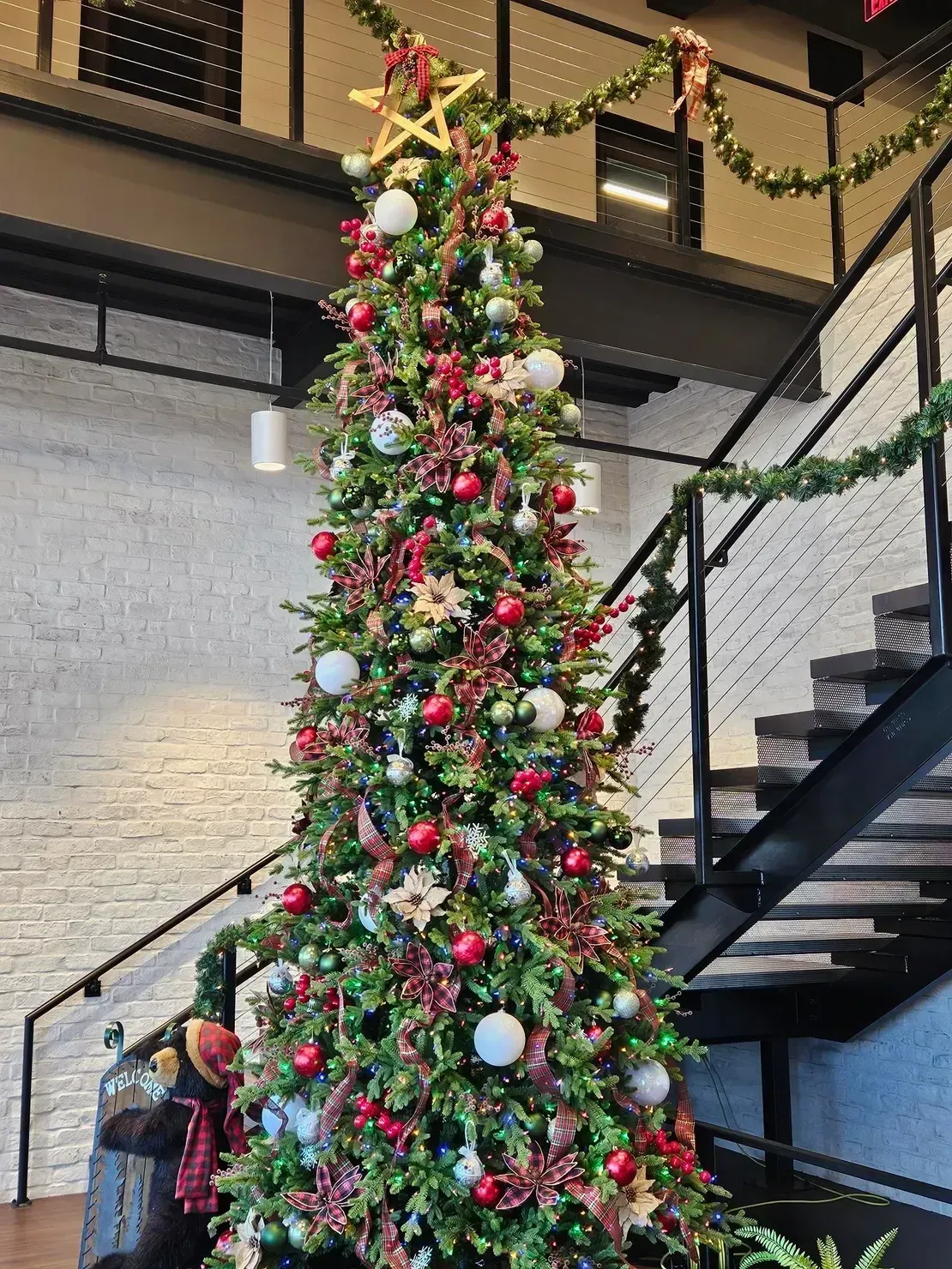 Tall, decorated Christmas tree with red and white ornaments and a gold star topper, standing next to a staircase.