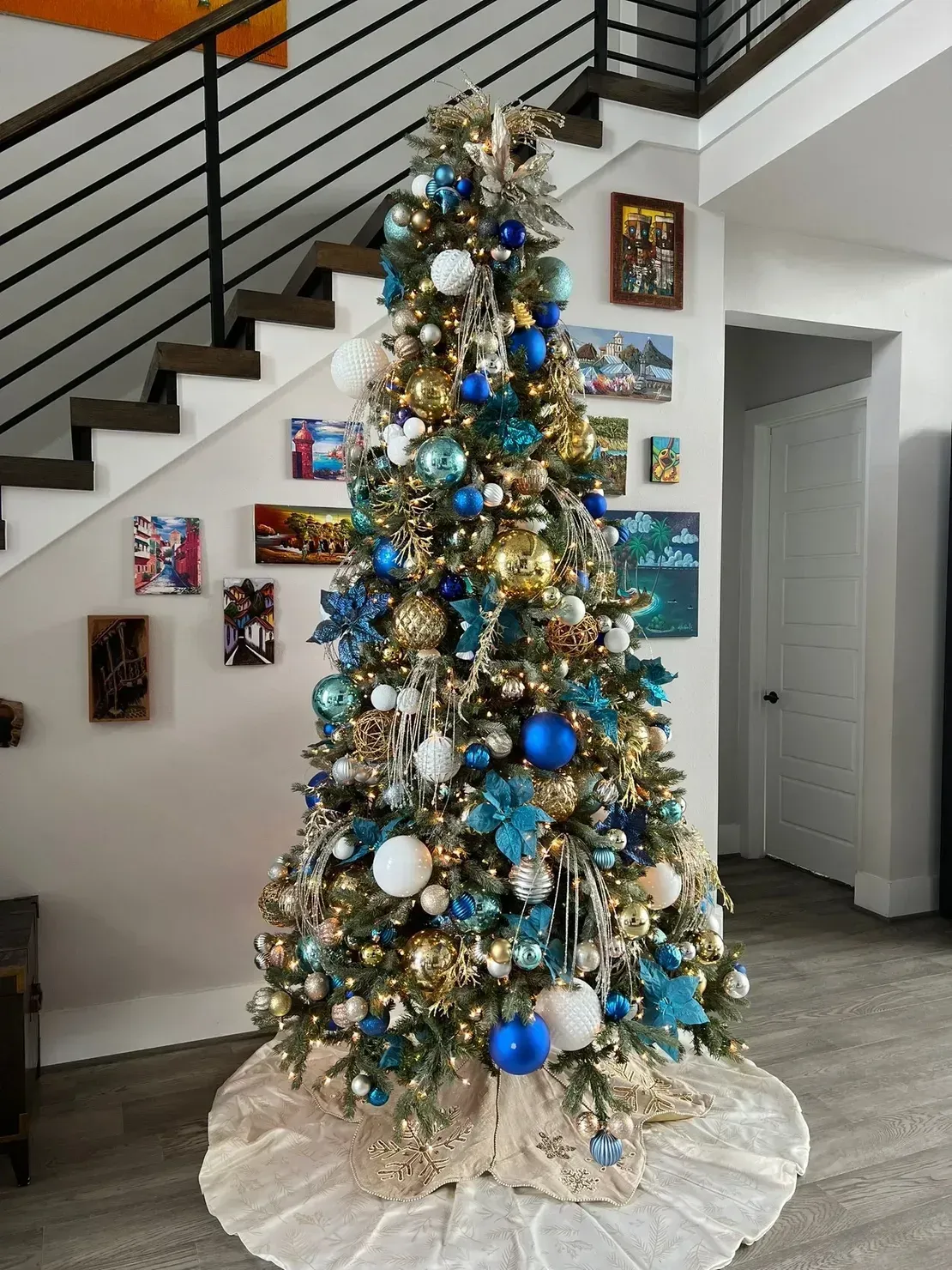 A tall Christmas tree decorated with blue, gold, and white ornaments, standing on a light fabric skirt in a home entry.