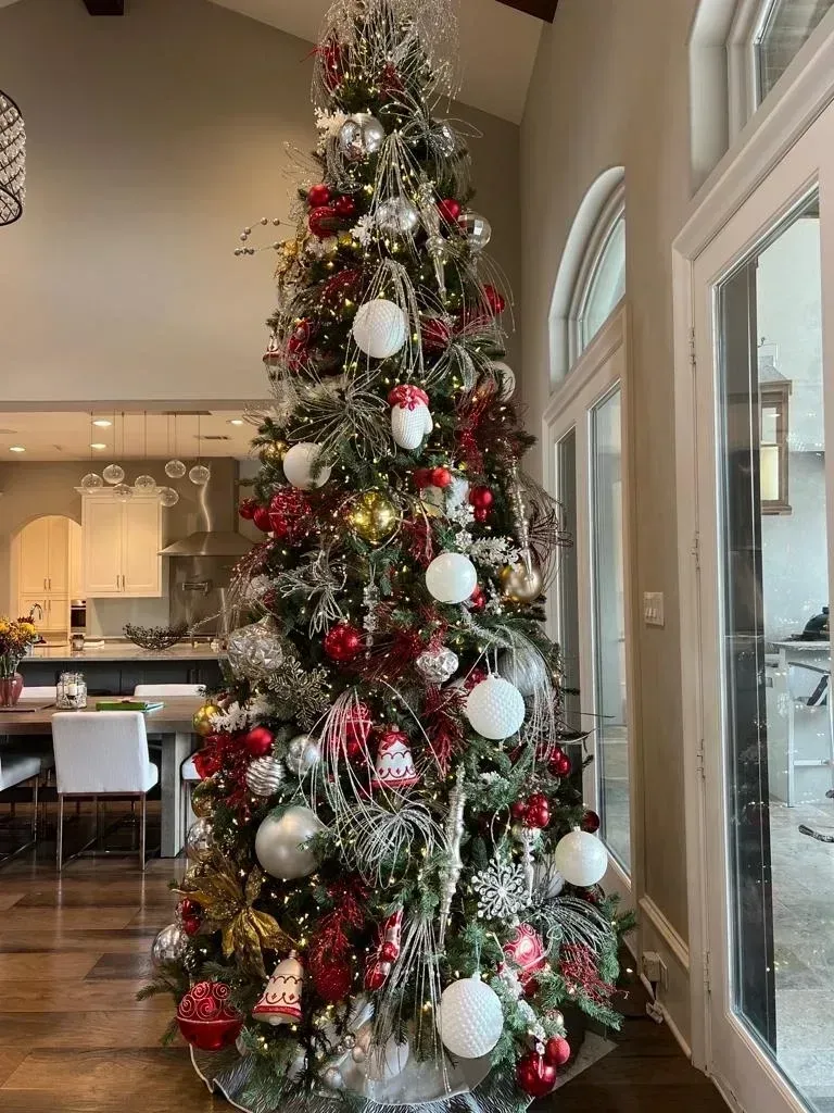 A tall, flocked Christmas tree decorated with red, silver, and gold ornaments stands in a bright, modern living room.