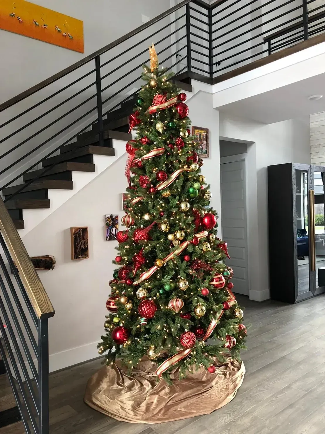 A tall Christmas tree decorated with red and gold ornaments and ribbons, standing in a room with a staircase.