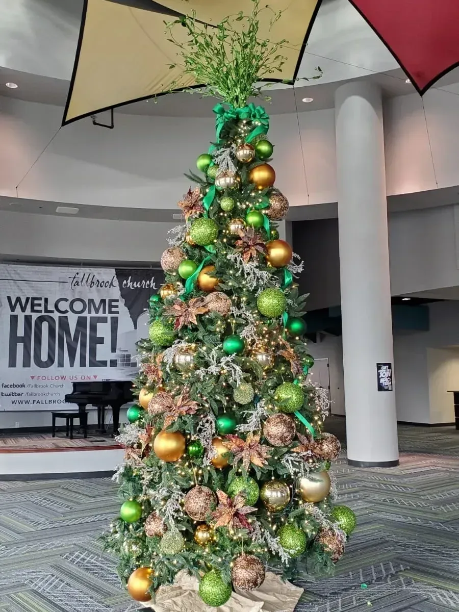 A tall Christmas tree decorated with gold and green ornaments stands in a lobby with a 