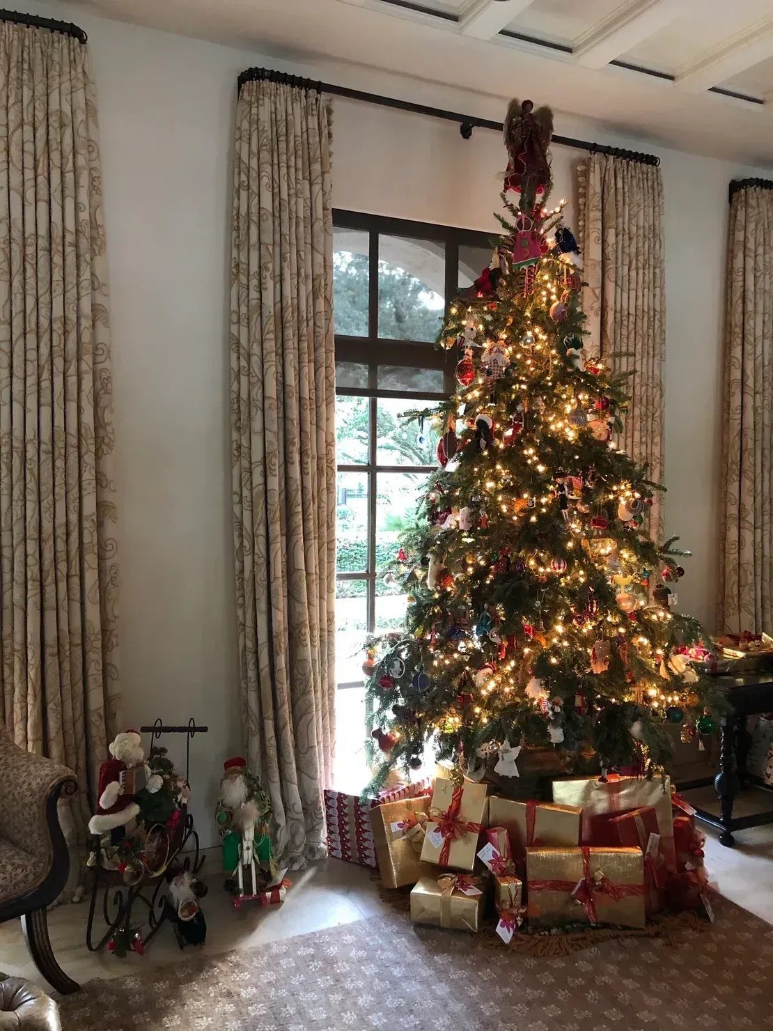 A lit Christmas tree decorated with ornaments stands in a room with tall patterned curtains and wrapped gifts at its base.