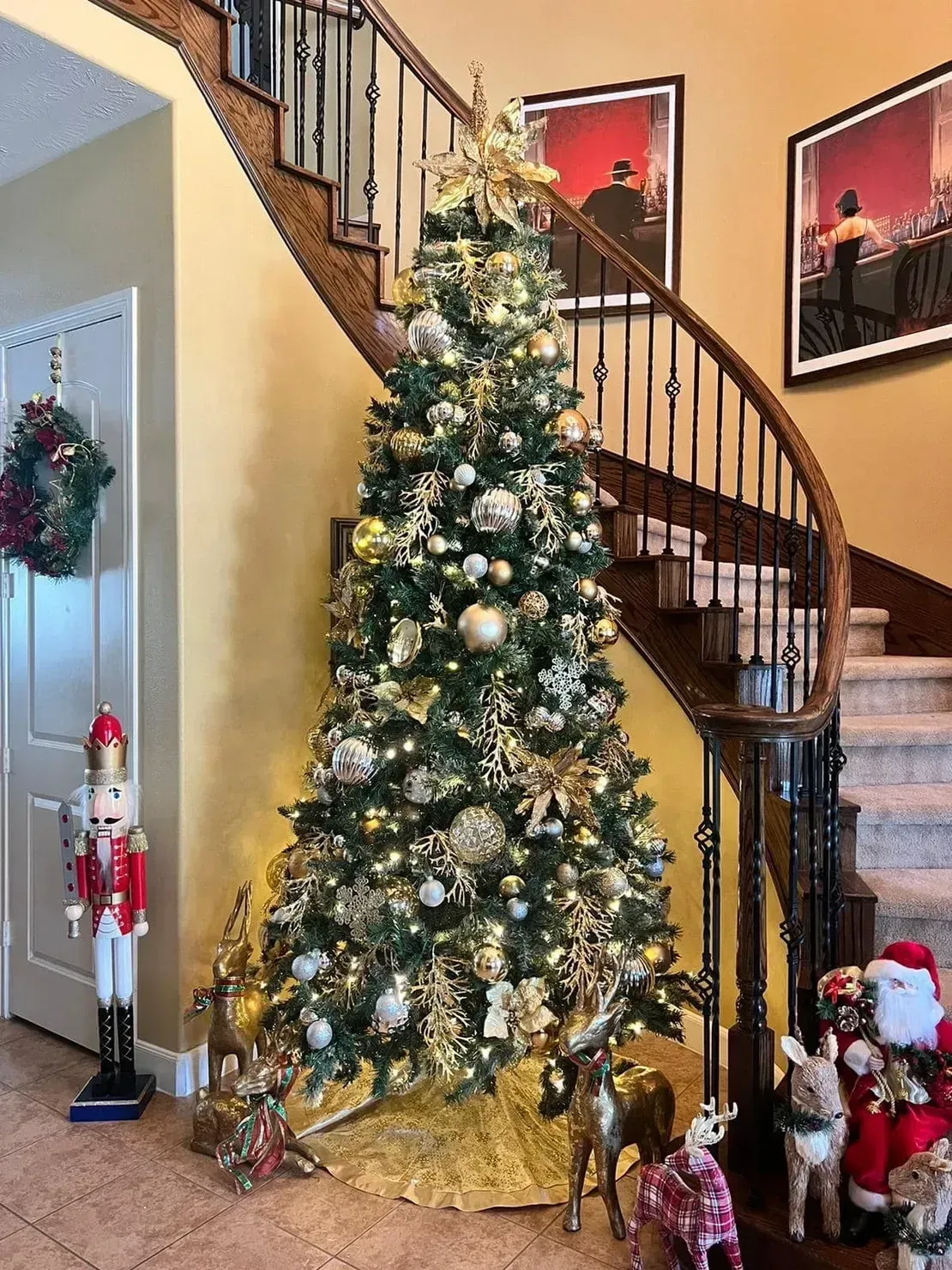 A tall Christmas tree decorated with gold and white ornaments, standing in a foyer with a staircase and festive figures.