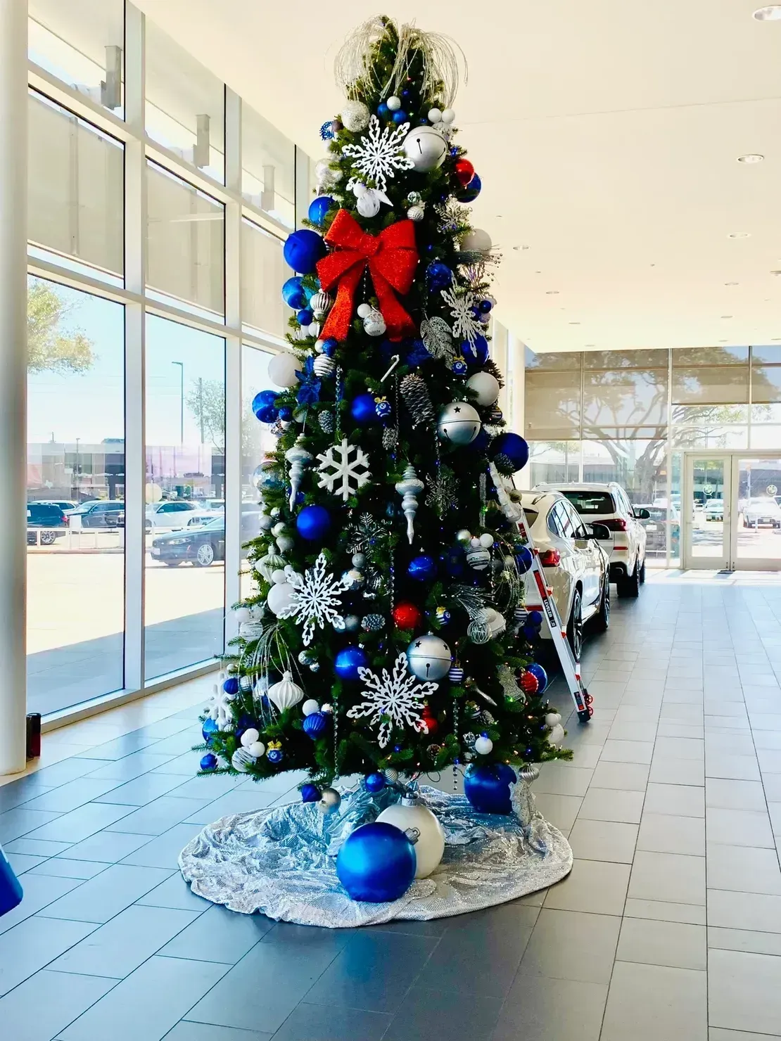 A tall Christmas tree decorated with blue, silver, and red ornaments, a large red bow, and snowflakes, set in a showroom.