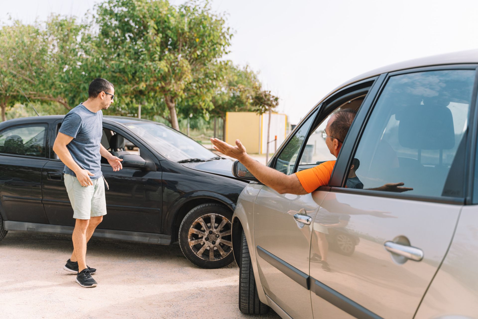 Man gestures at a car after an accident. Driver in the other car gestures back. Scene is outdoors.