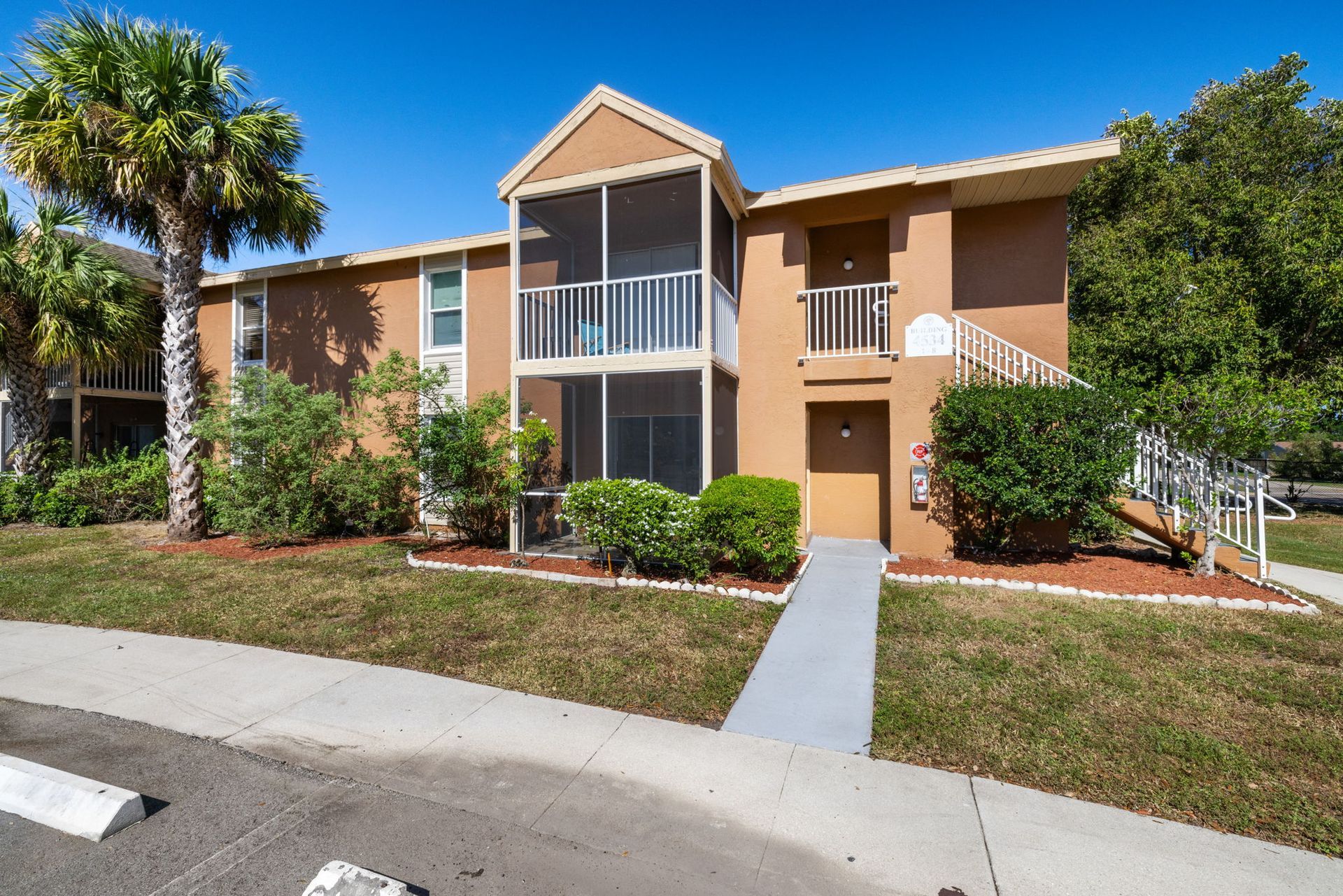 Tan apartment building with a screened balcony, accessible ramp, and palm tree in front.