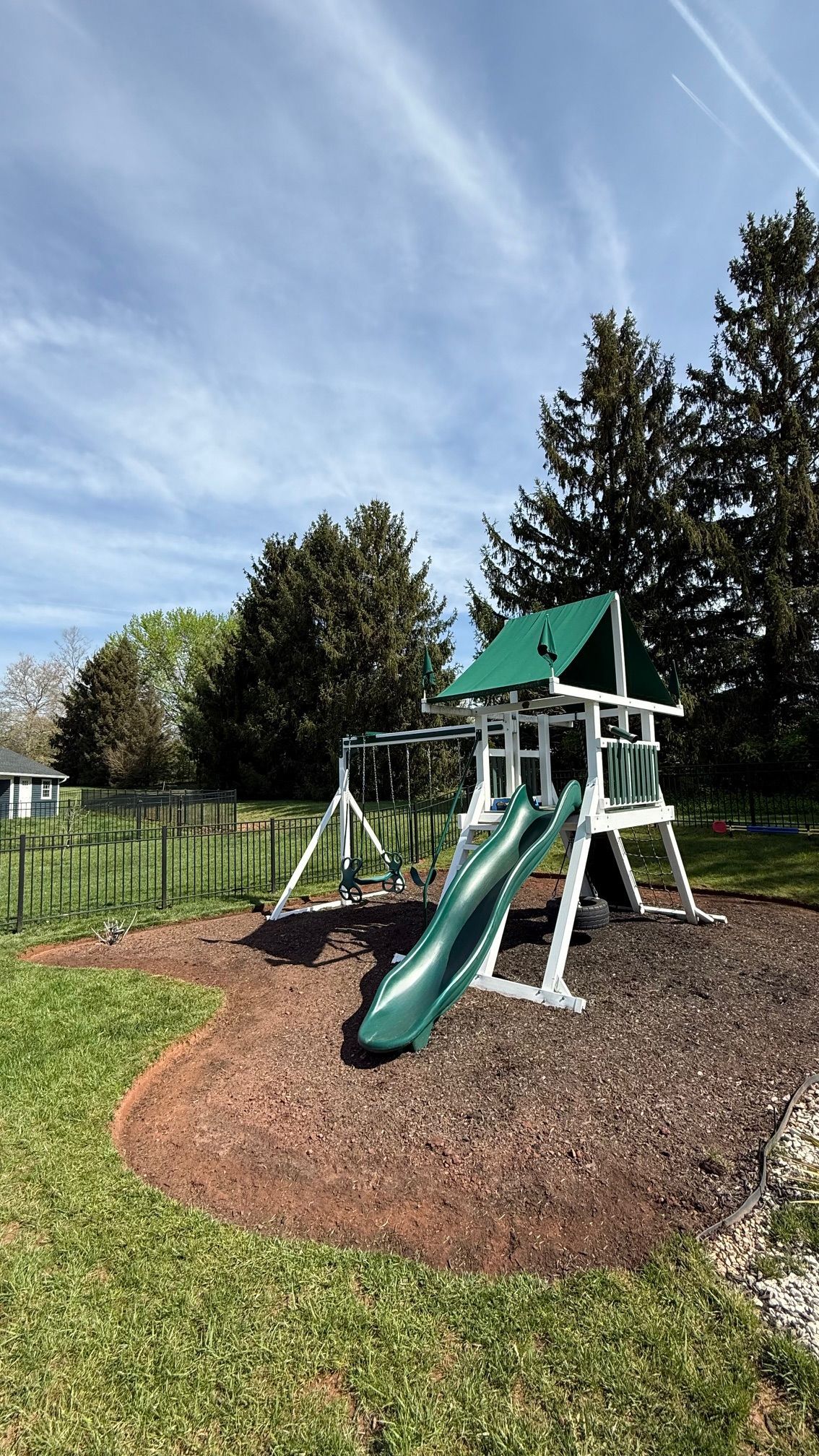 Playground with a green slide, swings, and a wooden playhouse on a mulch bed, surrounded by trees and grass.