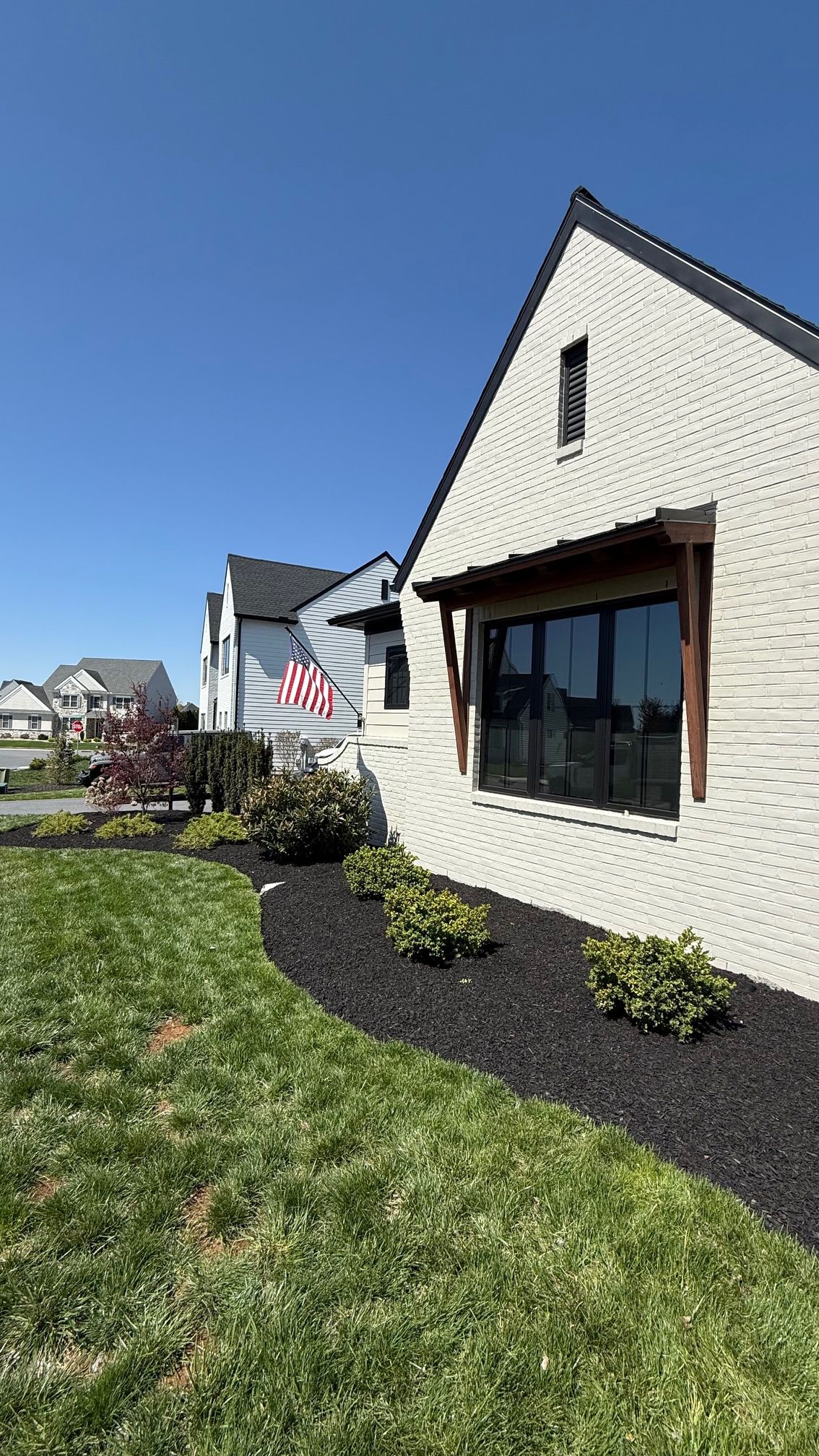 Beige brick house with black trim, a large window, and American flag on a sunny day.