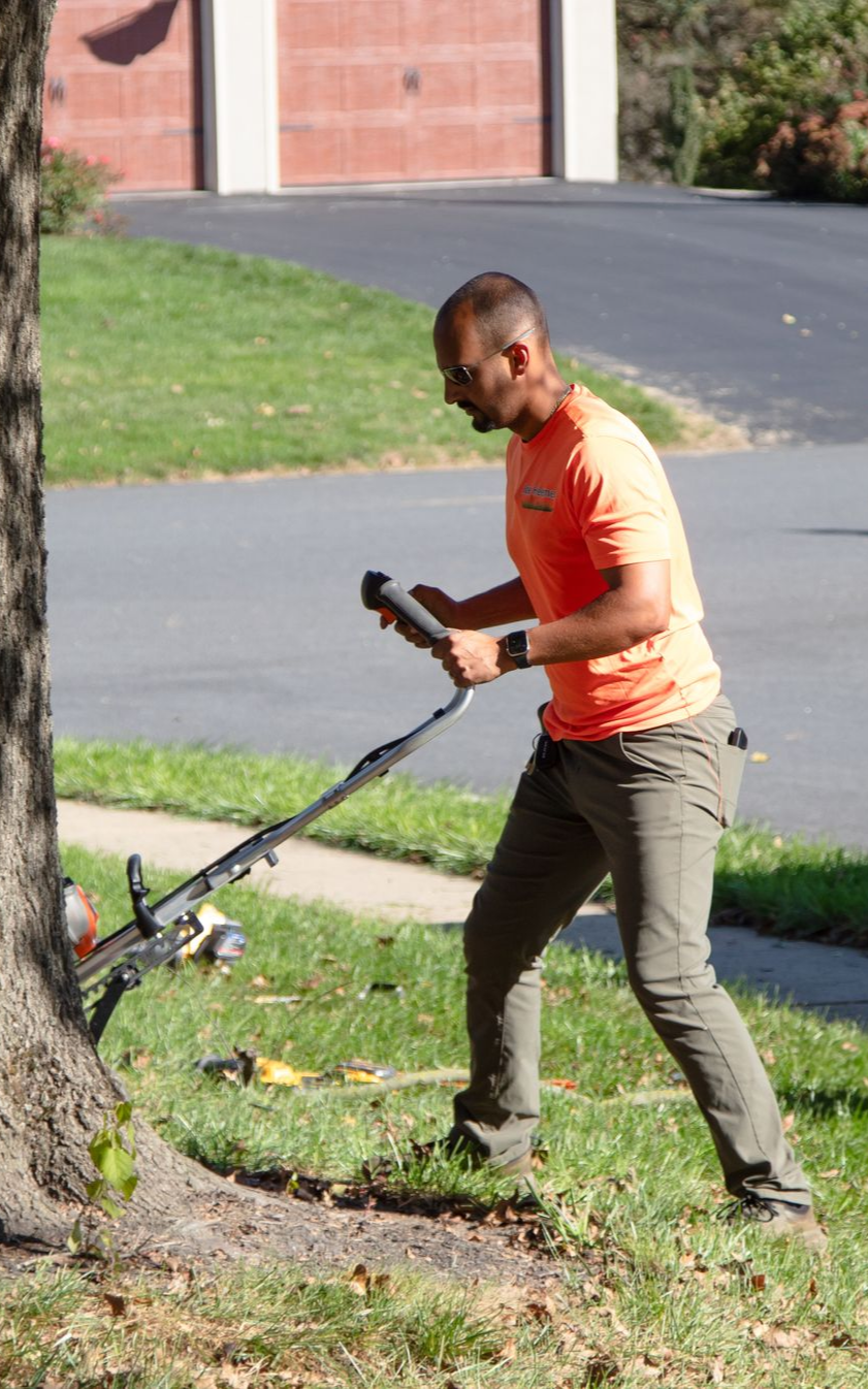 Person trimming grass near a tree with a trimmer. Wearing orange shirt, brown pants, in a grassy yard.