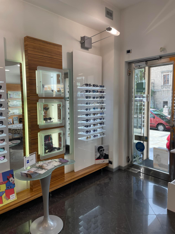 Interno di un negozio di ottica con occhiali in esposizione. Porta aperta, pavimento scuro e illuminazione intensa.