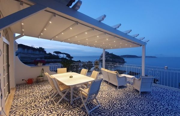 Patio con mobili bianchi sotto una tenda da sole retrattile, con vista sul mare al tramonto.