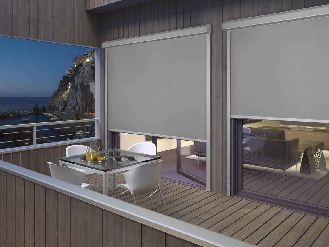 Balcone con tende grigie parzialmente abbassate, tavolo con sedie, vista serale sull'acqua e sulle scogliere.