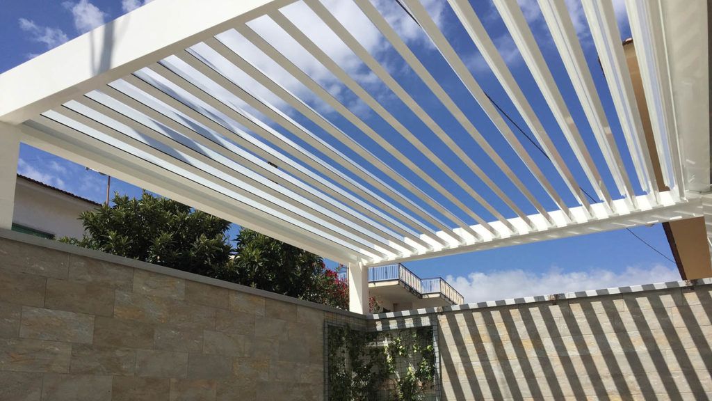 Tetto bianco del pergolato con stecche angolate contro un cielo azzurro, sopra un muro di pietra.