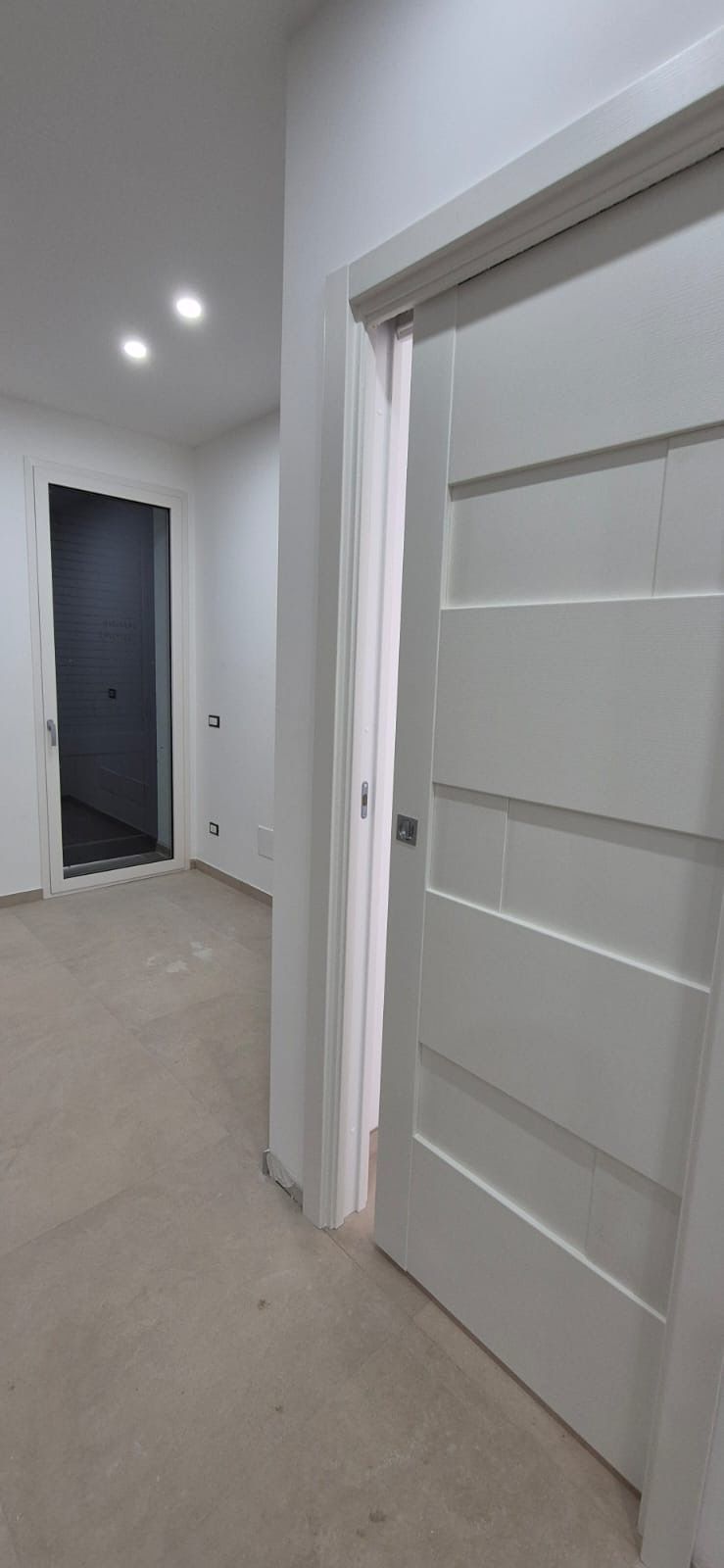 Corridoio interno con pareti bianche e una porta bianca; un'altra porta aperta è visibile in fondo al corridoio.