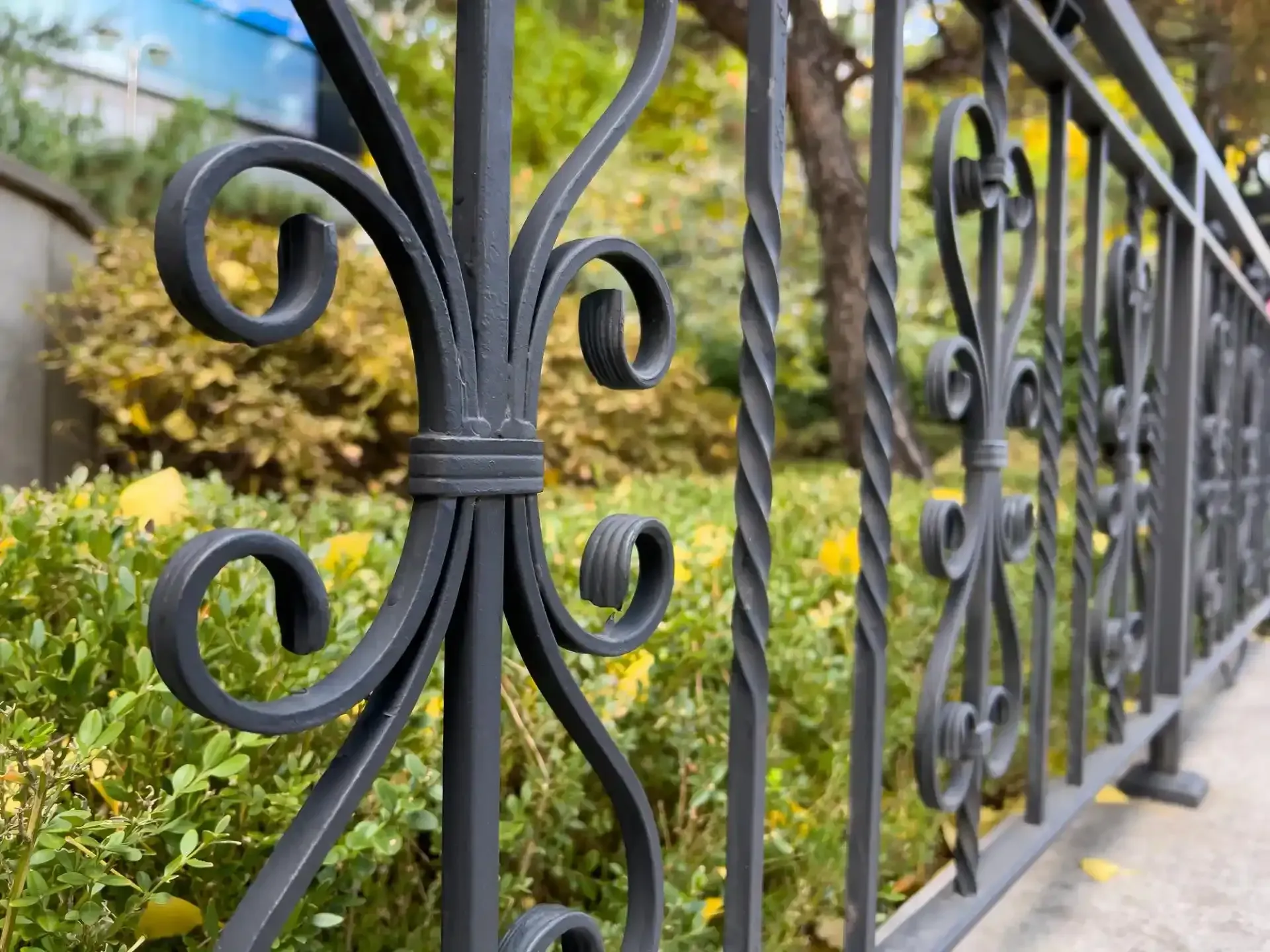 Primo piano di una recinzione decorativa in ferro battuto nero con vortici e un palo attorcigliato, con vegetazione sfocata sullo sfondo.
