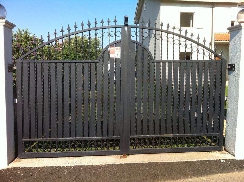 Cancelli d'accesso in metallo nero con parte superiore ad arco decorativa, inseriti in colonne di pietra bianca di fronte a una casa.