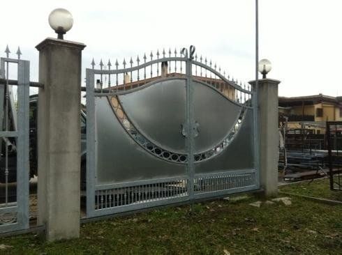 Cancello metallico con motivo decorativo, fiancheggiato da pilastri in cemento con luci a globo, di fronte a un edificio.