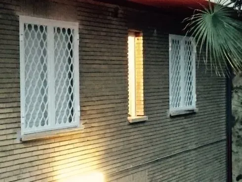 Facciata di un edificio in mattoni con tre finestre. Due hanno grate di sicurezza bianche, una è illuminata dall'interno. È visibile una foglia di palma.