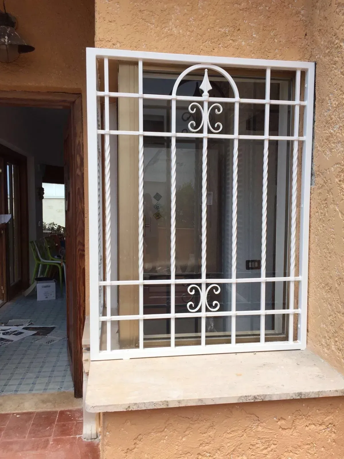 Inferriate di sicurezza bianche su una finestra in una parete di stucco arancione, accanto a una porta marrone.