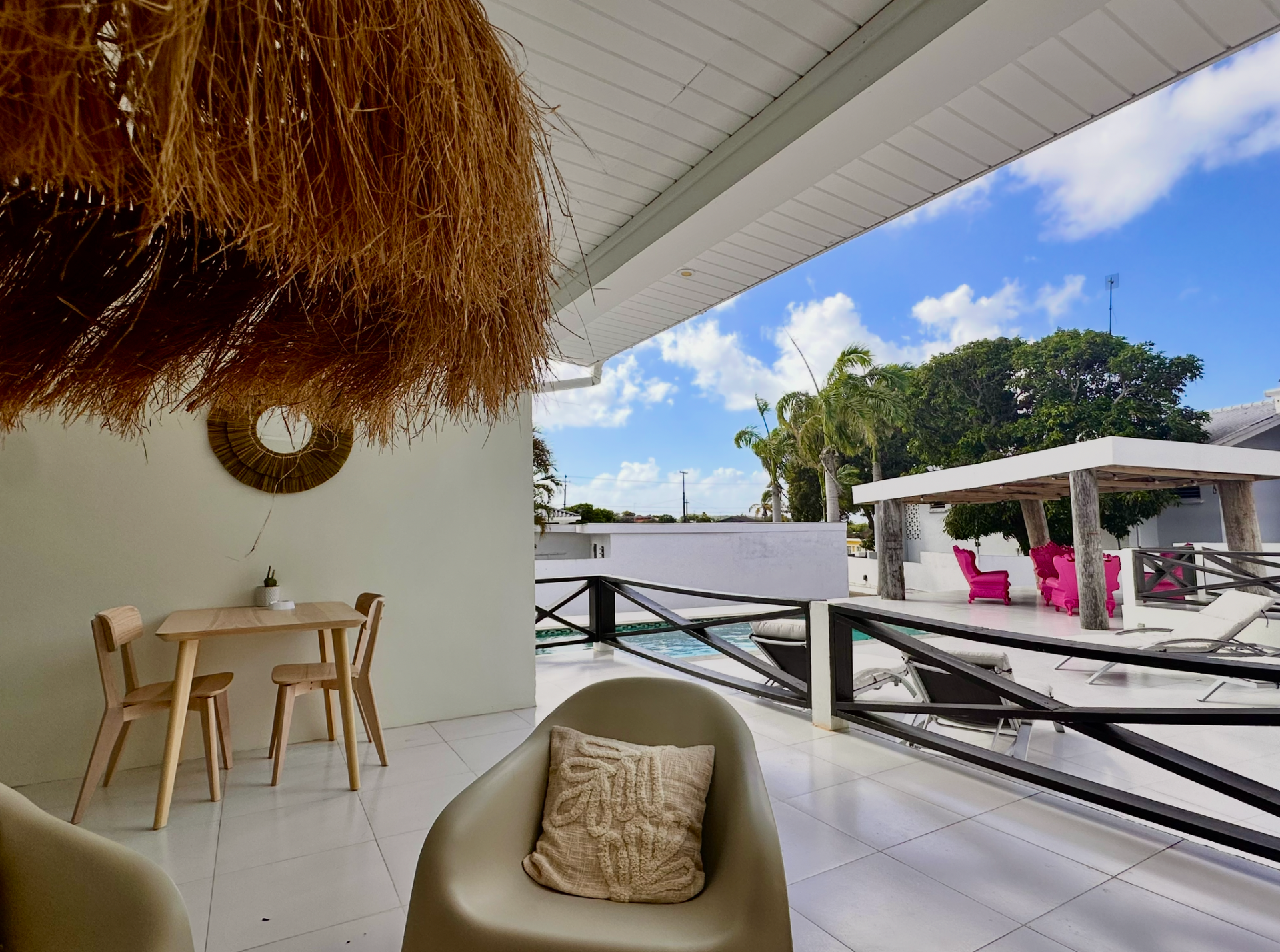 Patio with seating, table, and decorative straw hanging, overlooking a sunny outdoor area and blue sky.