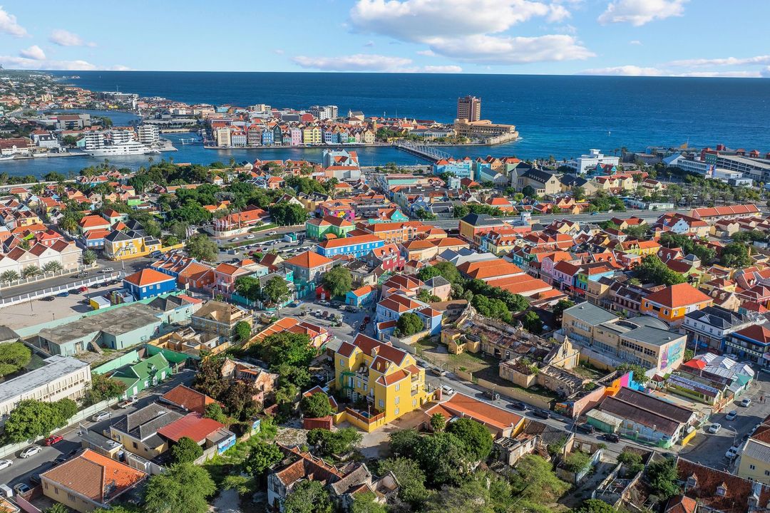 Aerial view of colorful buildings lining the waterfront in Willemstad, Curaçao, with the blue ocean in the background.