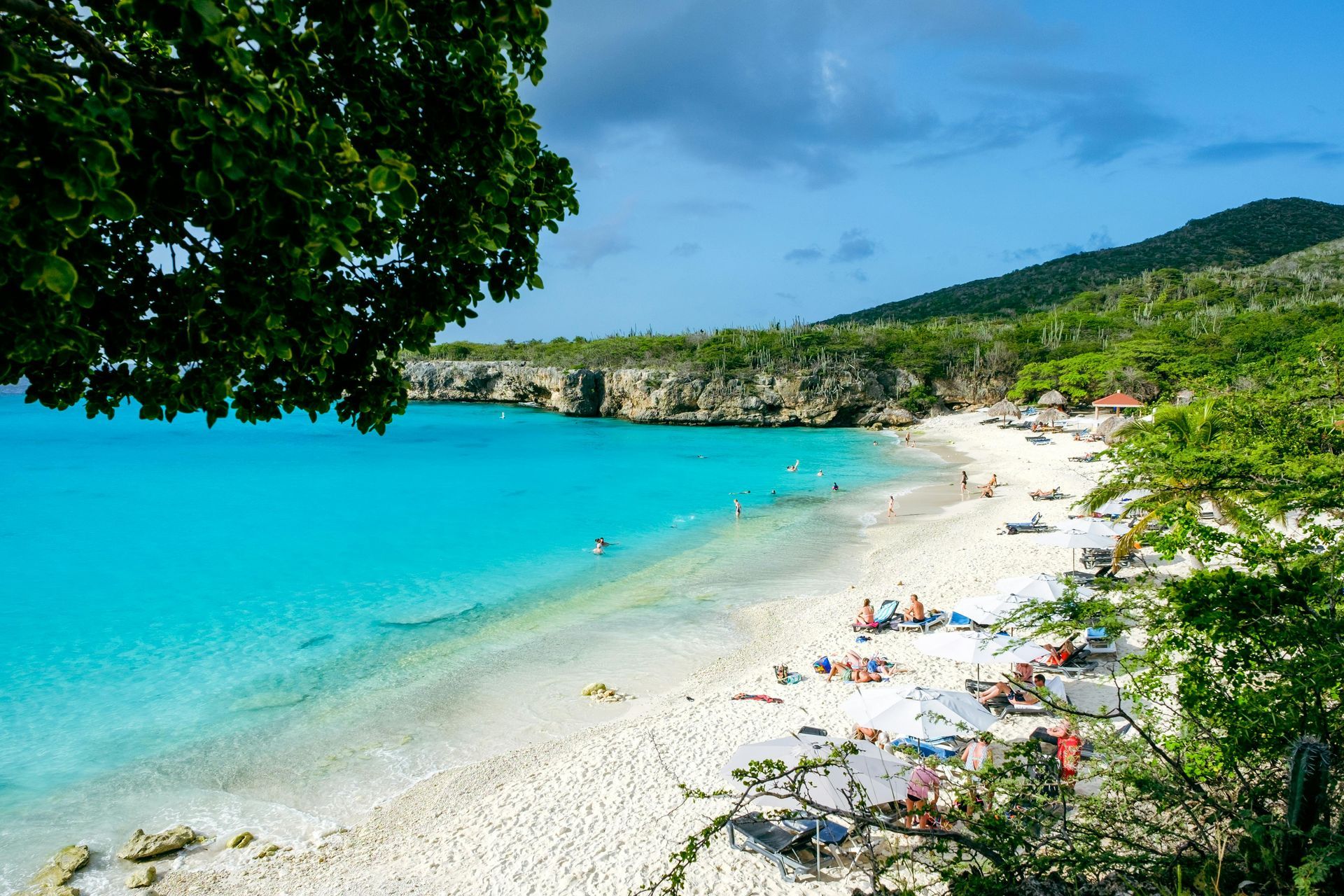 Beach with white sand, turquoise water, people, cliffs, and lush green vegetation under a blue sky.