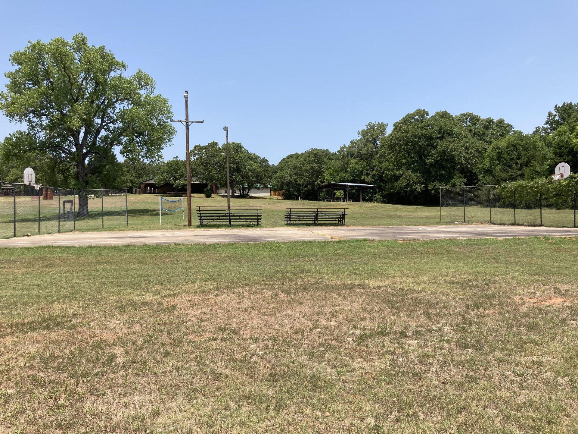 Grassy park with basketball hoops and two benches under a blue sky, trees in the background.