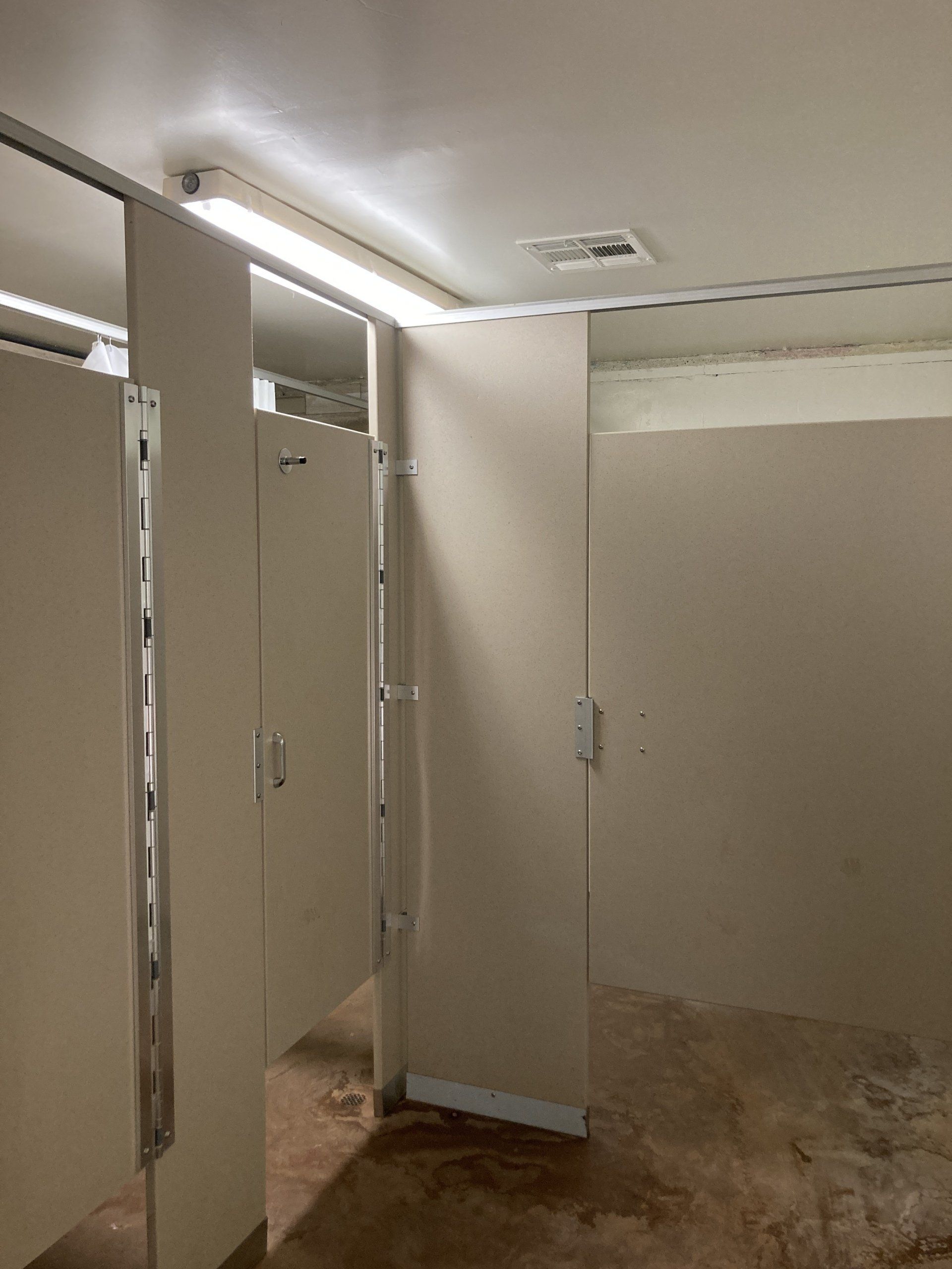 Empty public restroom with tan stalls and concrete floor. Fluorescent lights on ceiling.