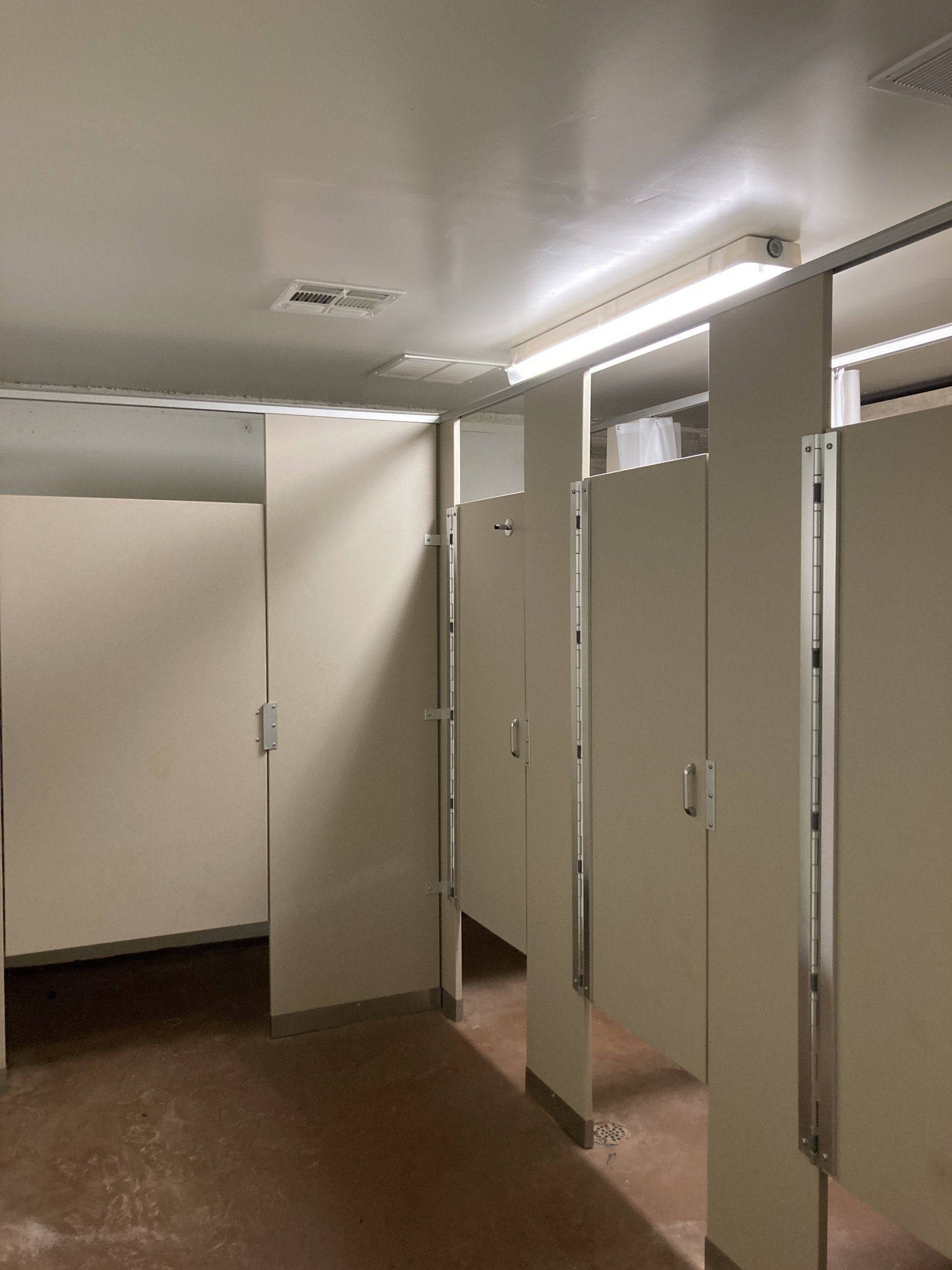 Empty public restroom with off-white stalls, light fixtures, and a reddish-brown floor.