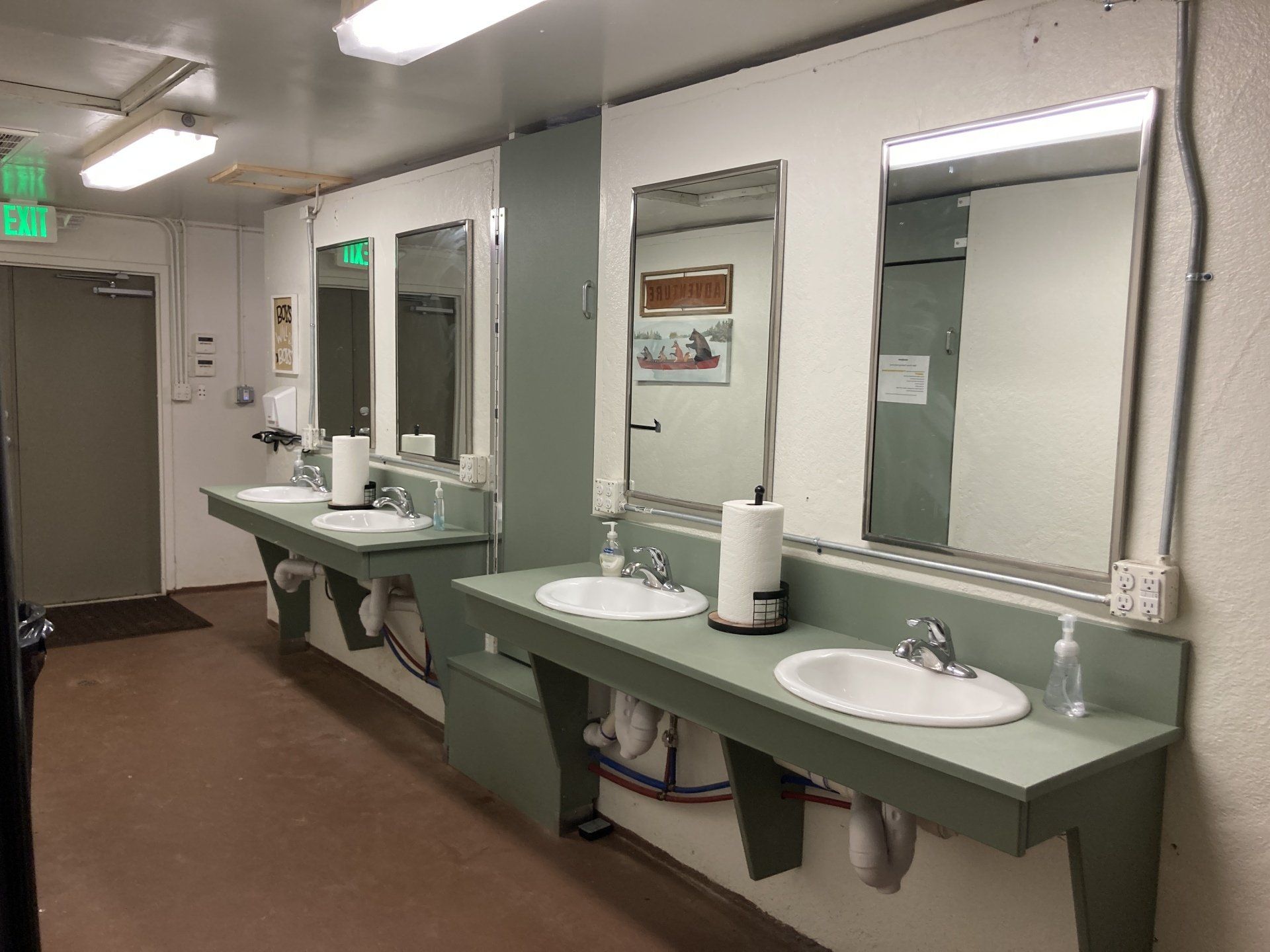 Public restroom with three sinks, mirrors, and paper towels, against a white wall and green counters.