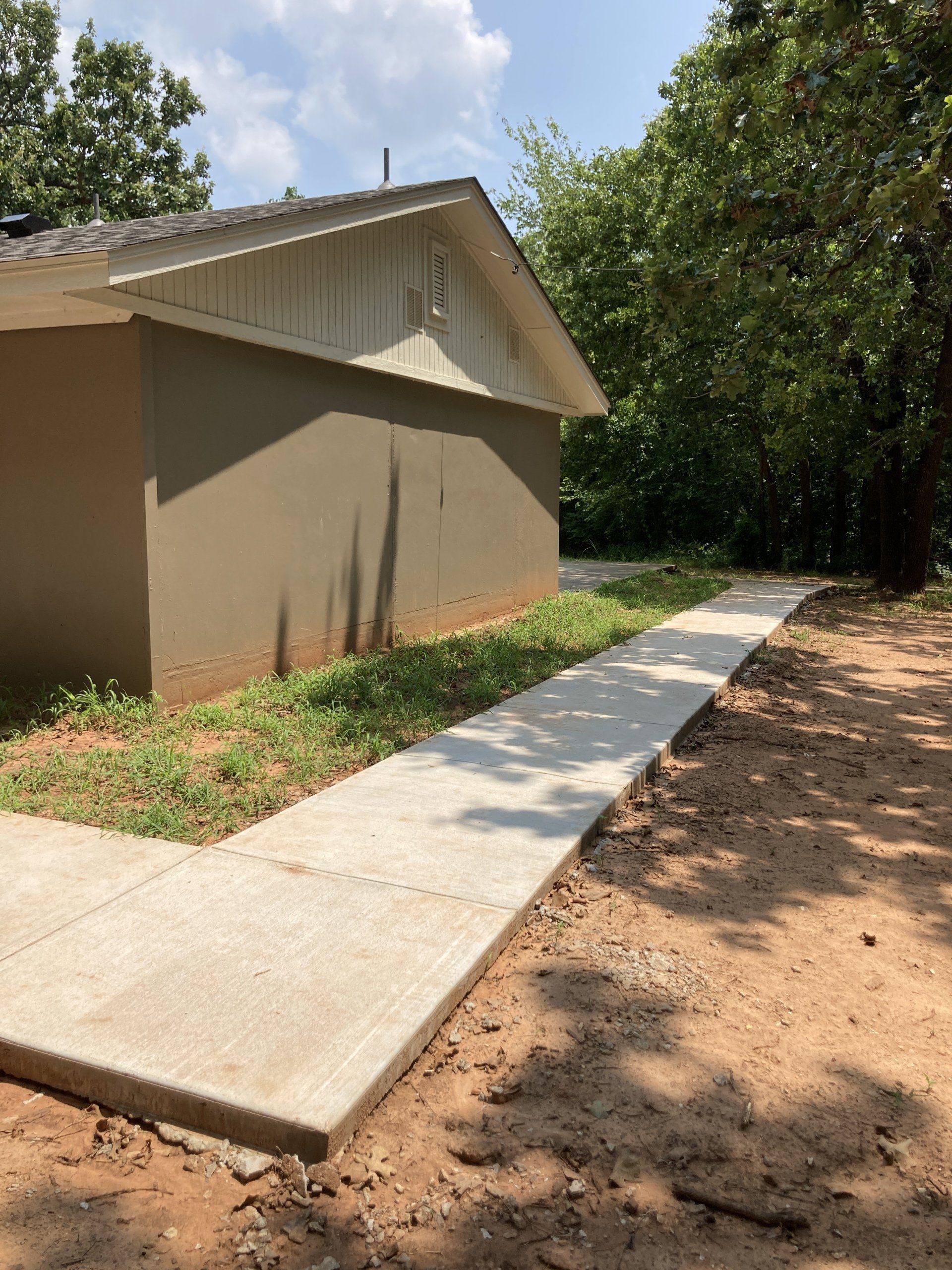 Concrete walkway beside a tan building with a sloped roof and surrounded by greenery on a sunny day.