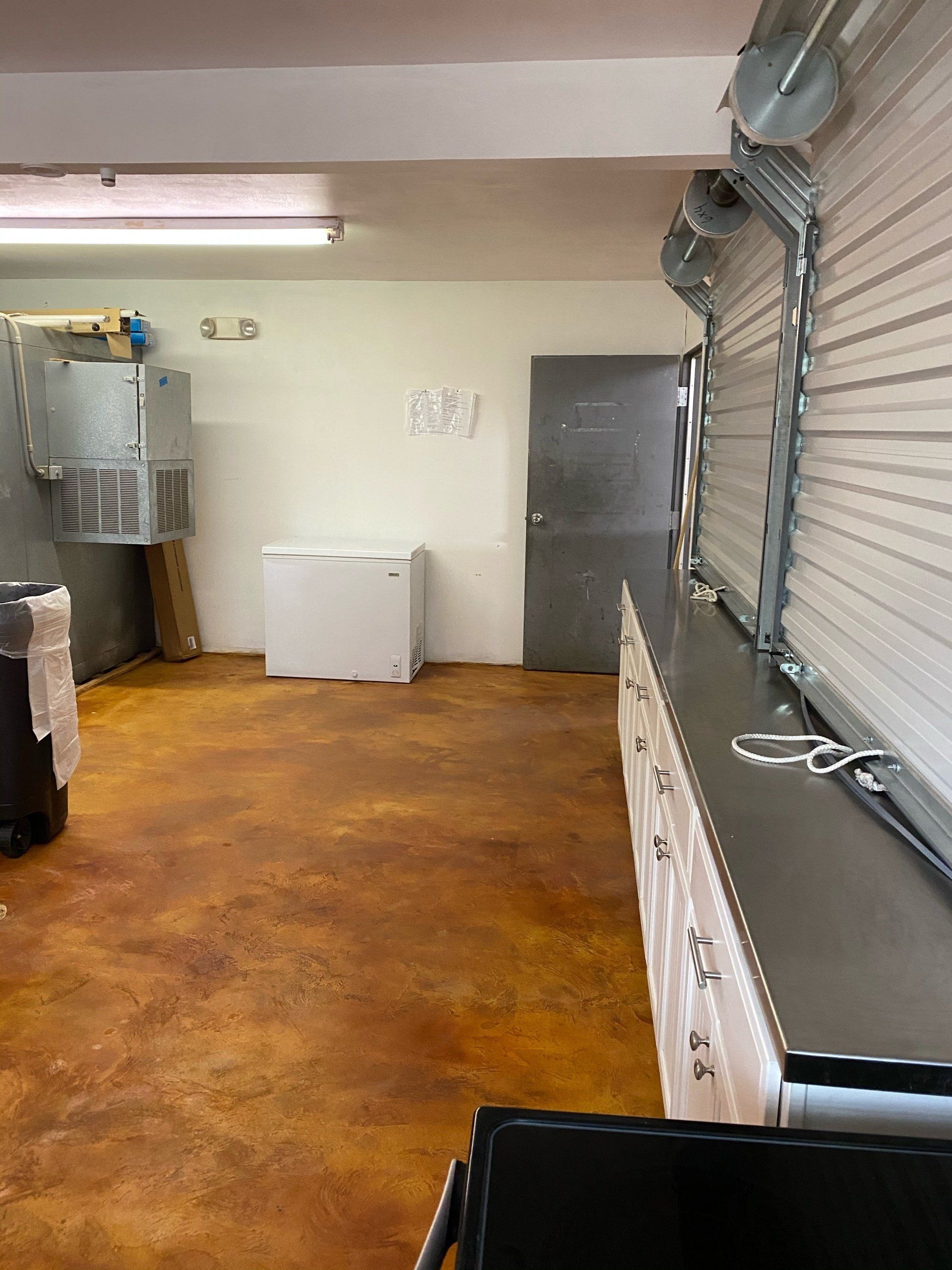 Interior view of a work space with a refrigerator, cabinets, a roll-up door, and stained concrete floor.