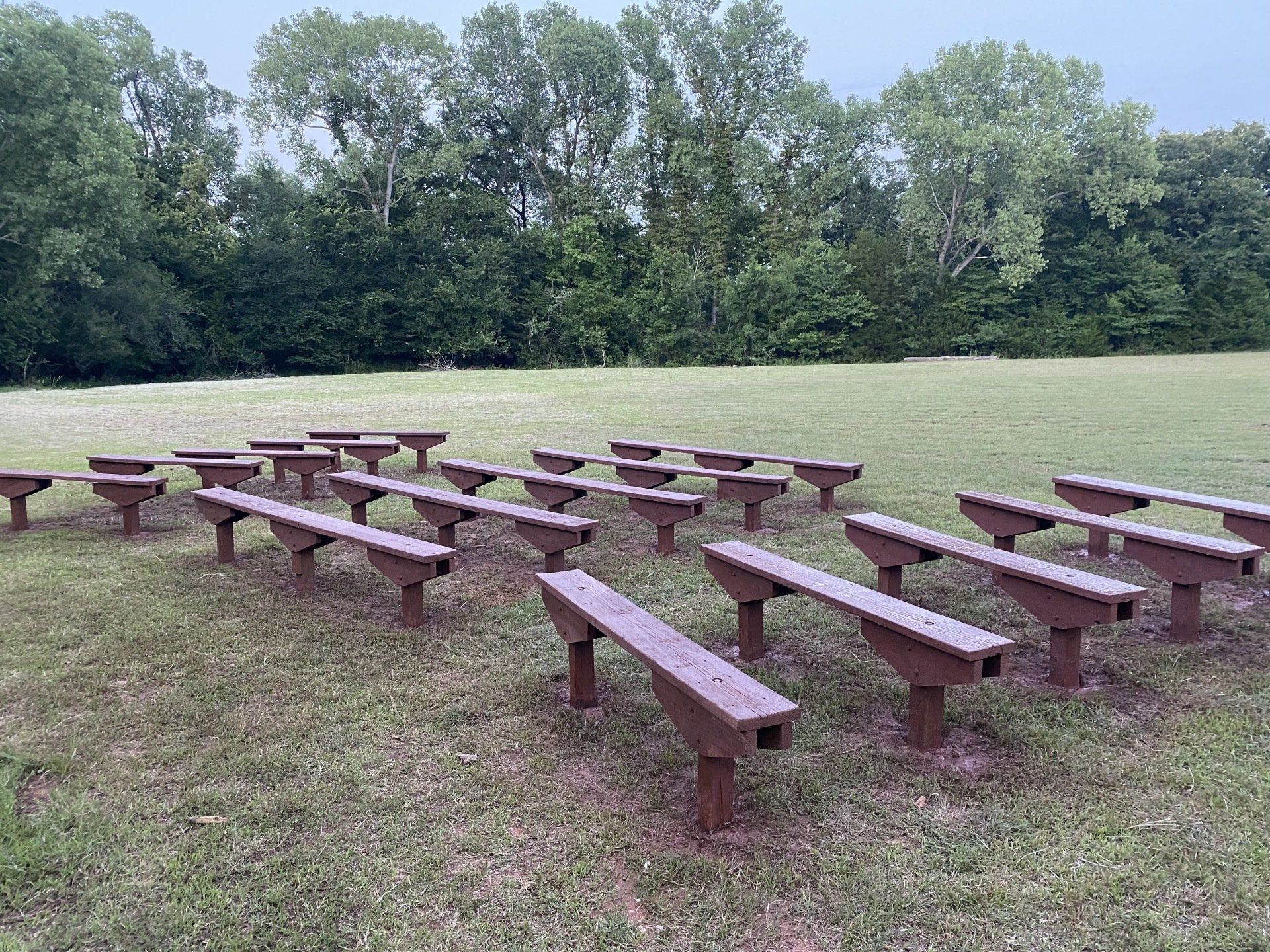Outdoor seating arrangement of brown benches on a grassy field, trees in the background.