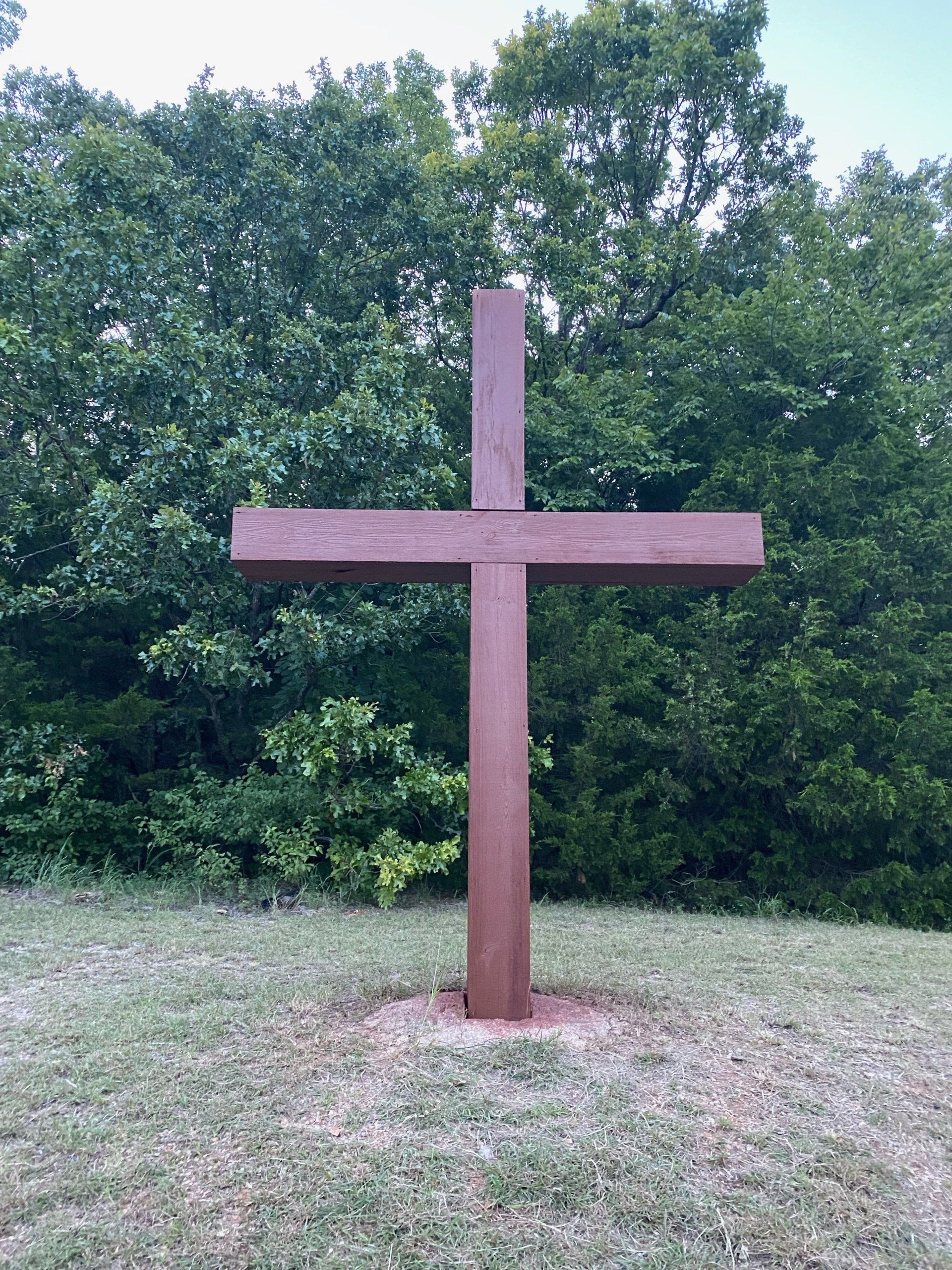 Wooden cross on a grassy hill, with a backdrop of green trees.