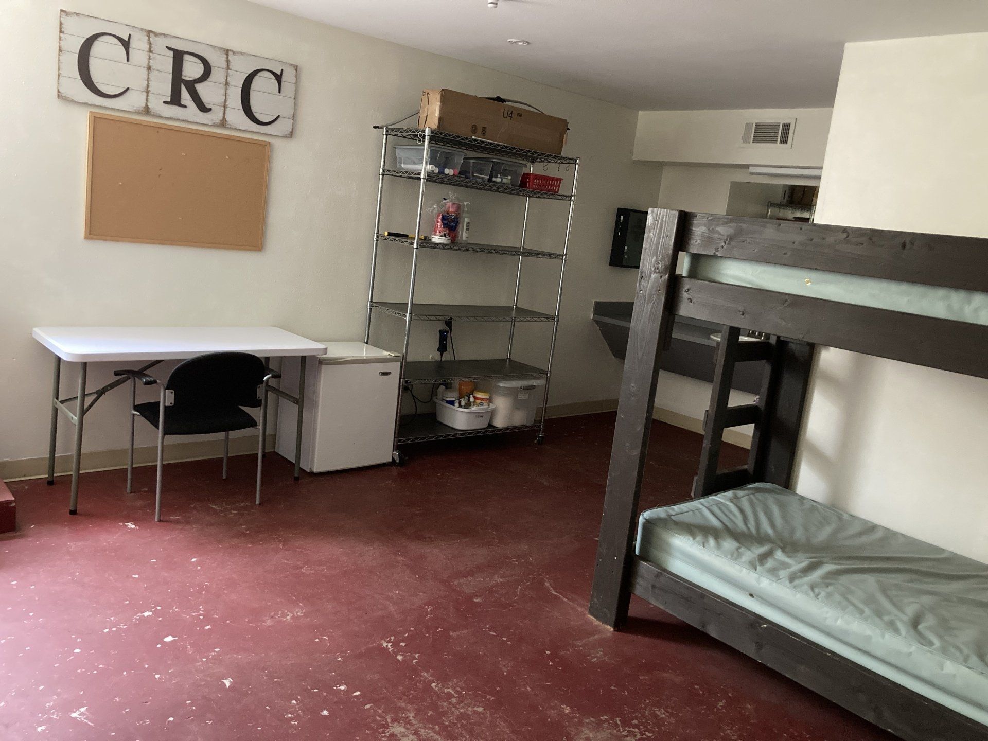 Room with bunk bed, desk, shelving unit, and mini-fridge. Red floor with white specks.