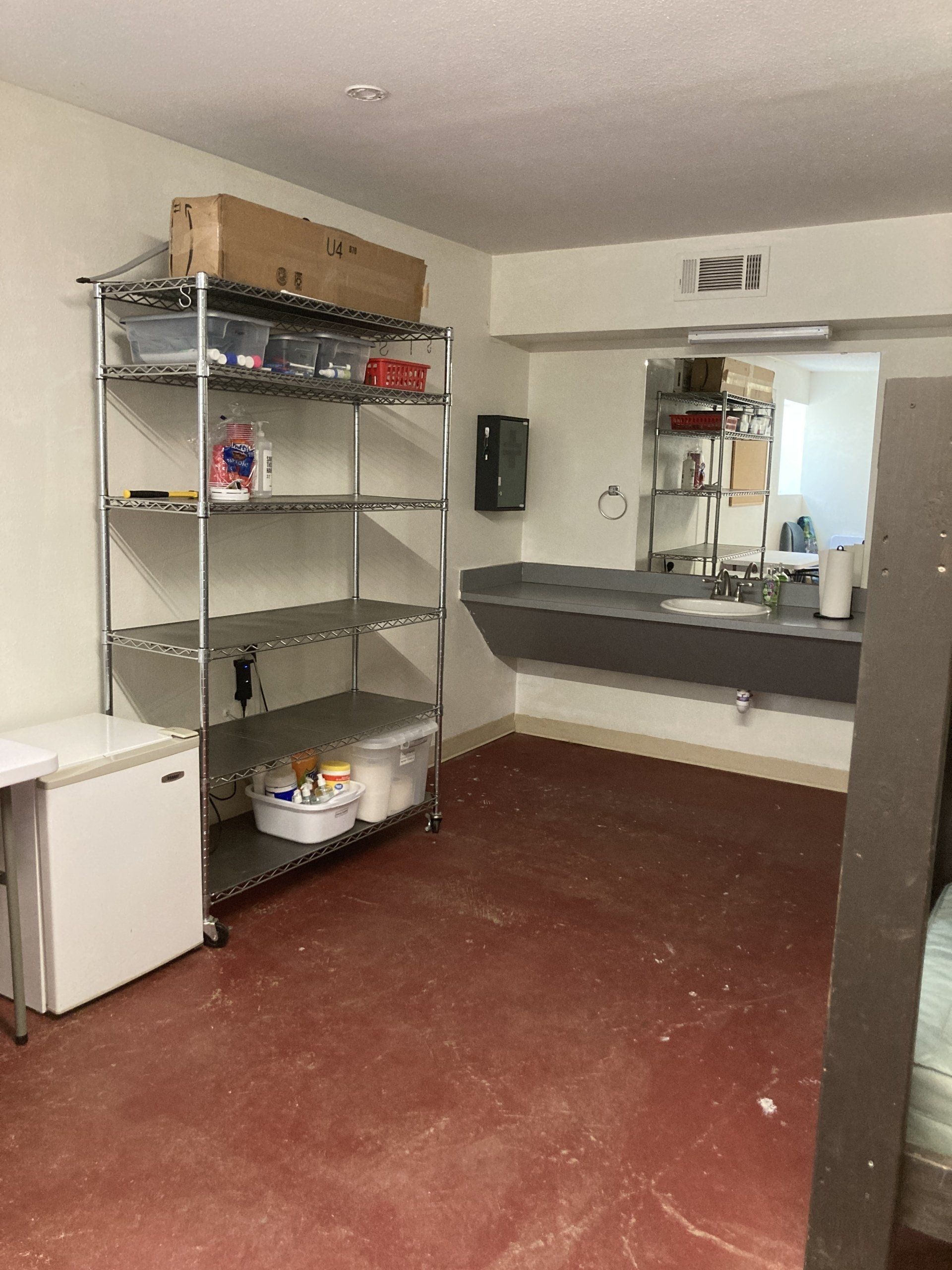 A storage room with a metal shelving unit, a sink, and a mini-fridge. Red floor.